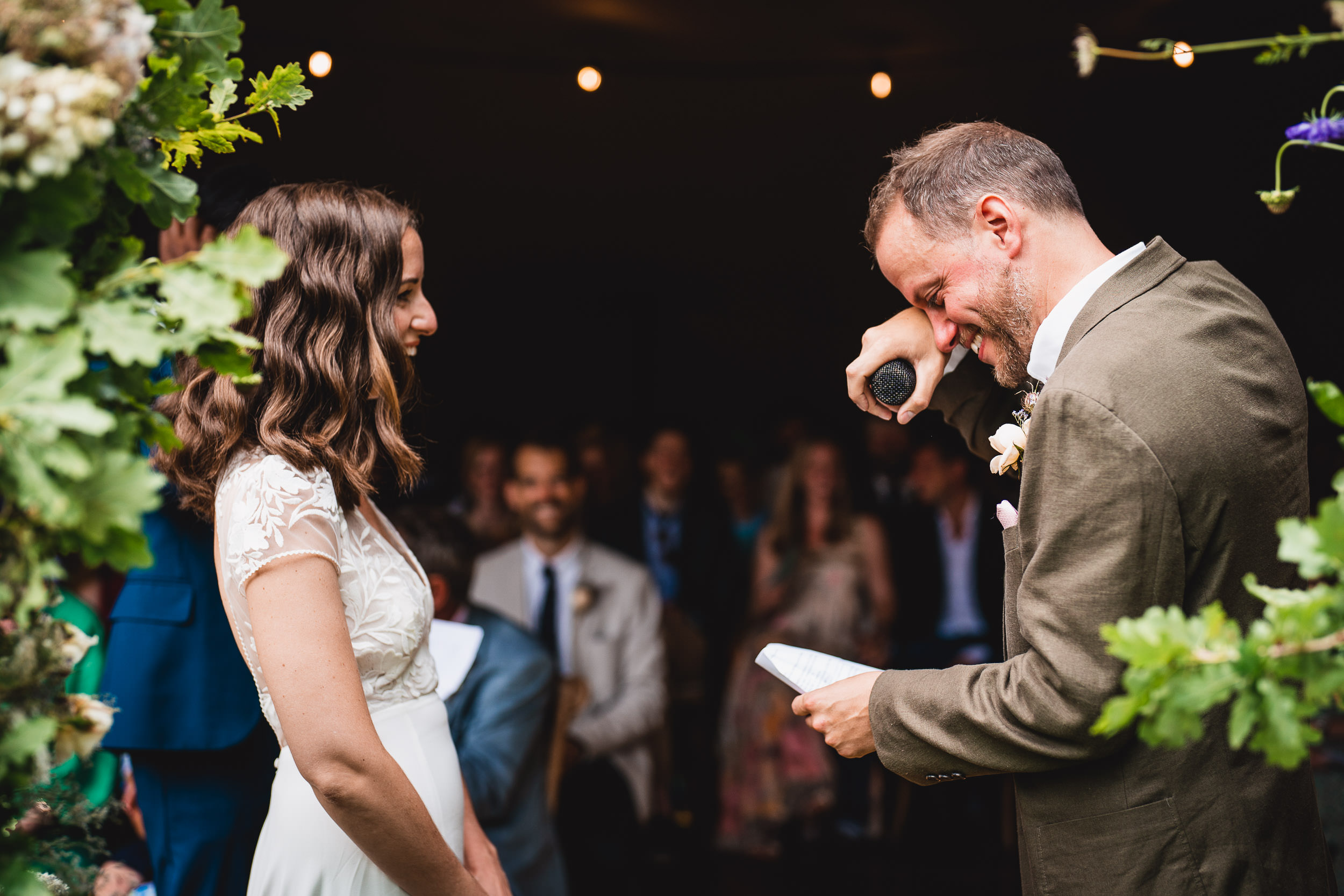 A man and a woman stand facing each other at a ceremony. The man holds a microphone and wipes his eye, while the woman smiles. Lush greenery frames the scene.