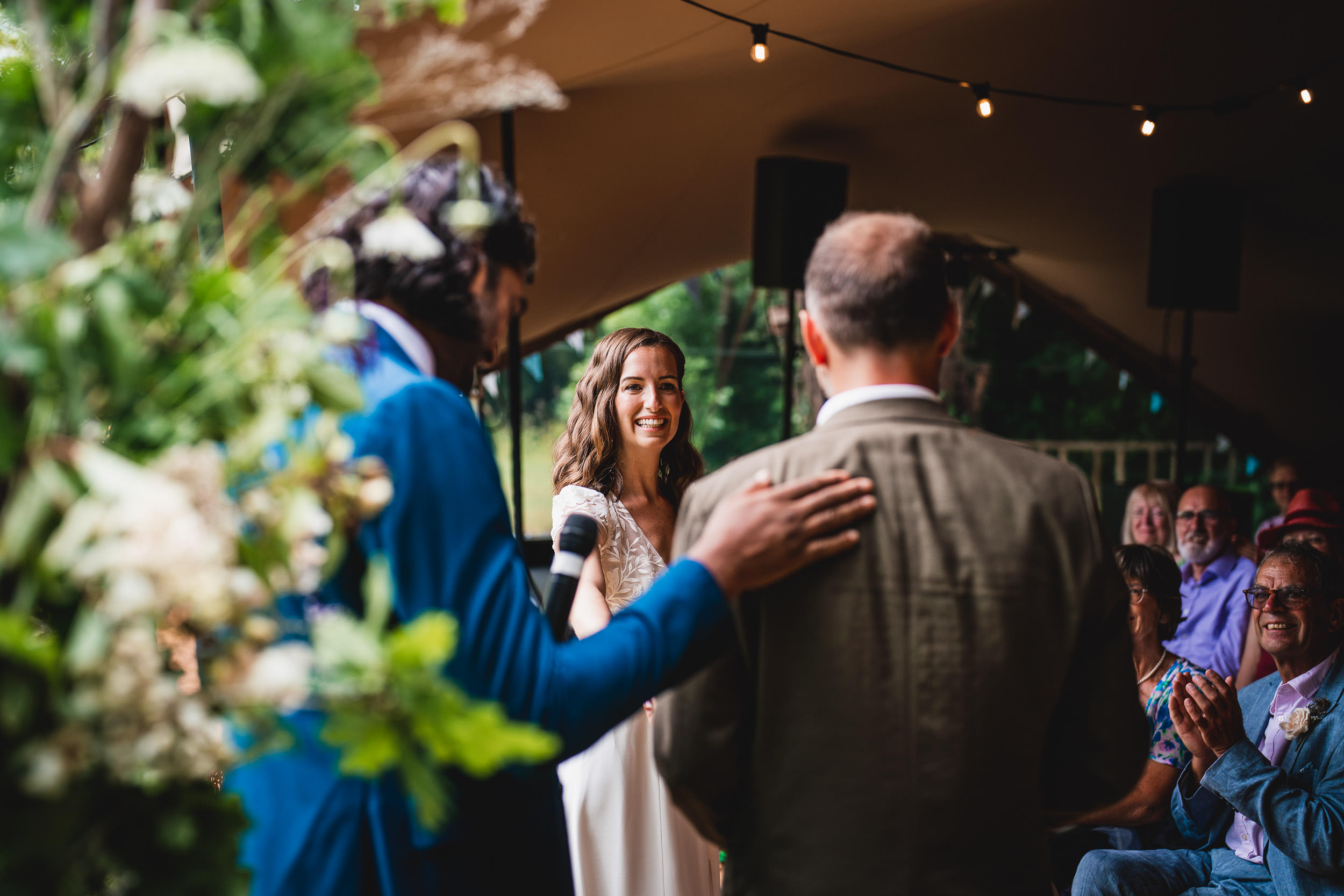 A bride smiles during a wedding ceremony. A man in a blue suit gestures with a microphone. Another man stands facing the bride. Guests are seated, clapping, and surrounded by greenery.