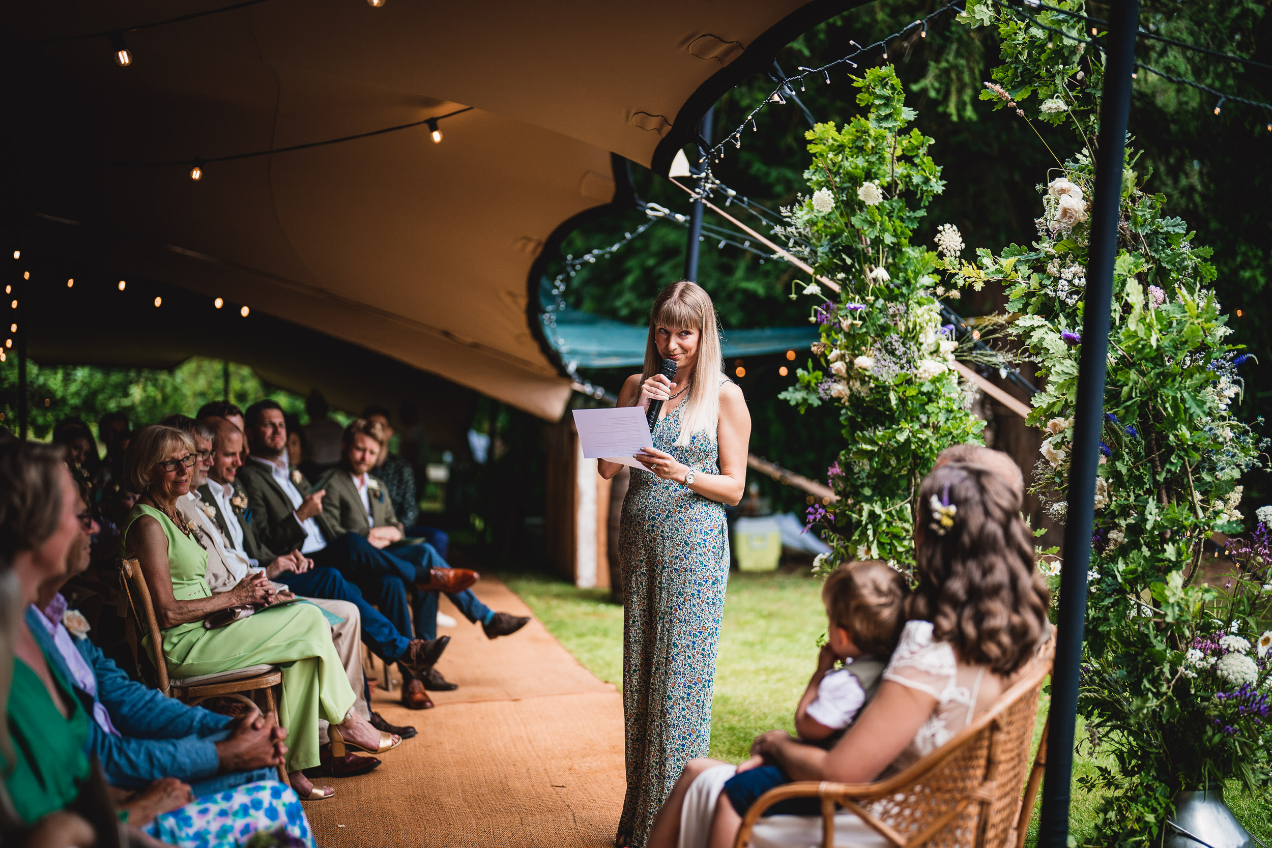 A woman reads from a paper at an outdoor event under a tent, while people seated listen. Flowers and greenery decorate the space.