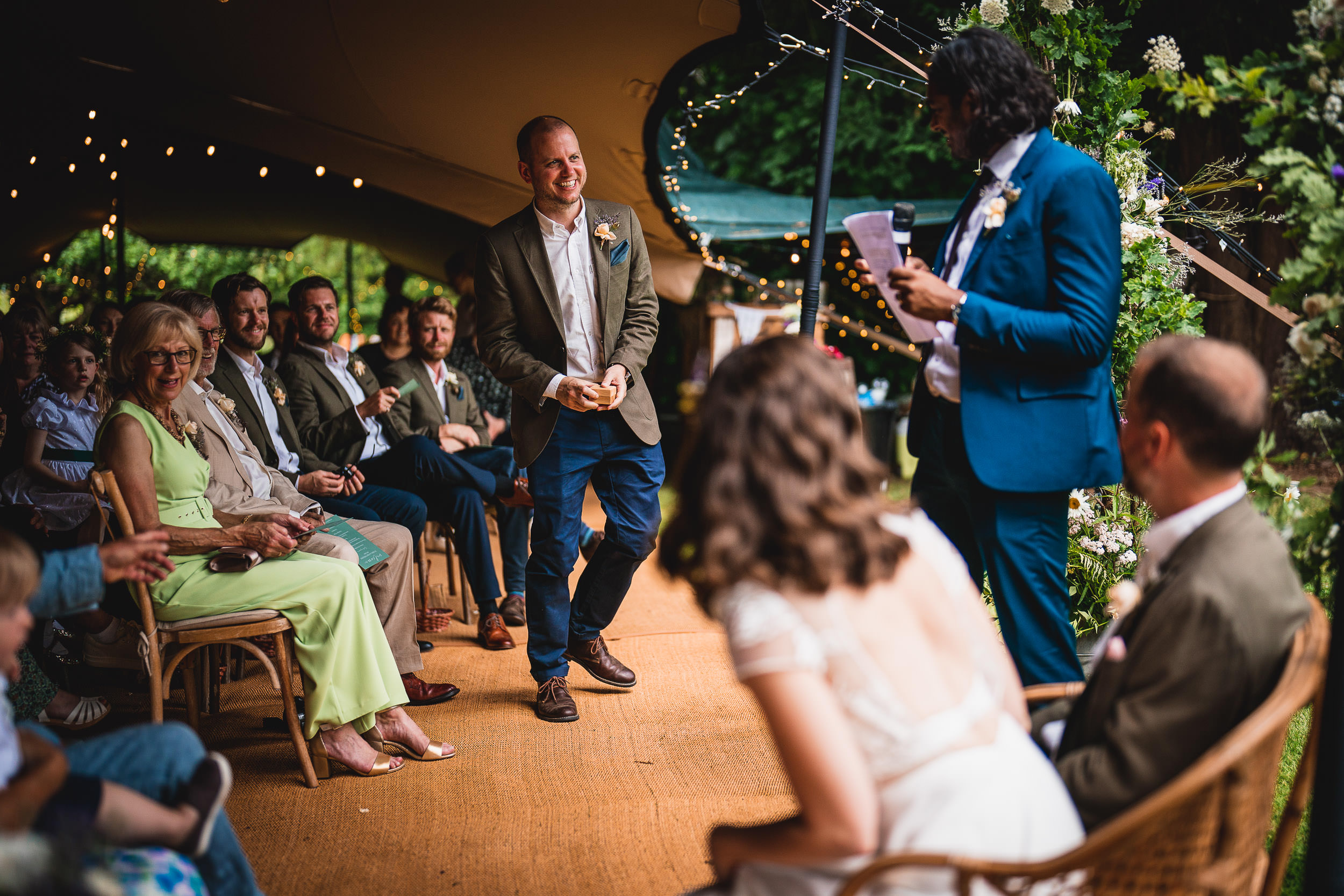 A man in a suit stands reading from a paper to a seated audience at an outdoor event, with string lights and greenery in the background.