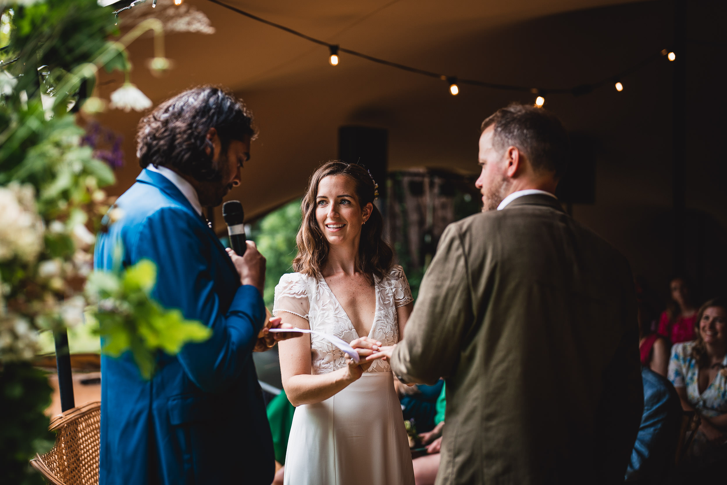 A couple exchanges vows during their wedding ceremony, with a person officiating nearby. The setting has string lights and floral decorations.