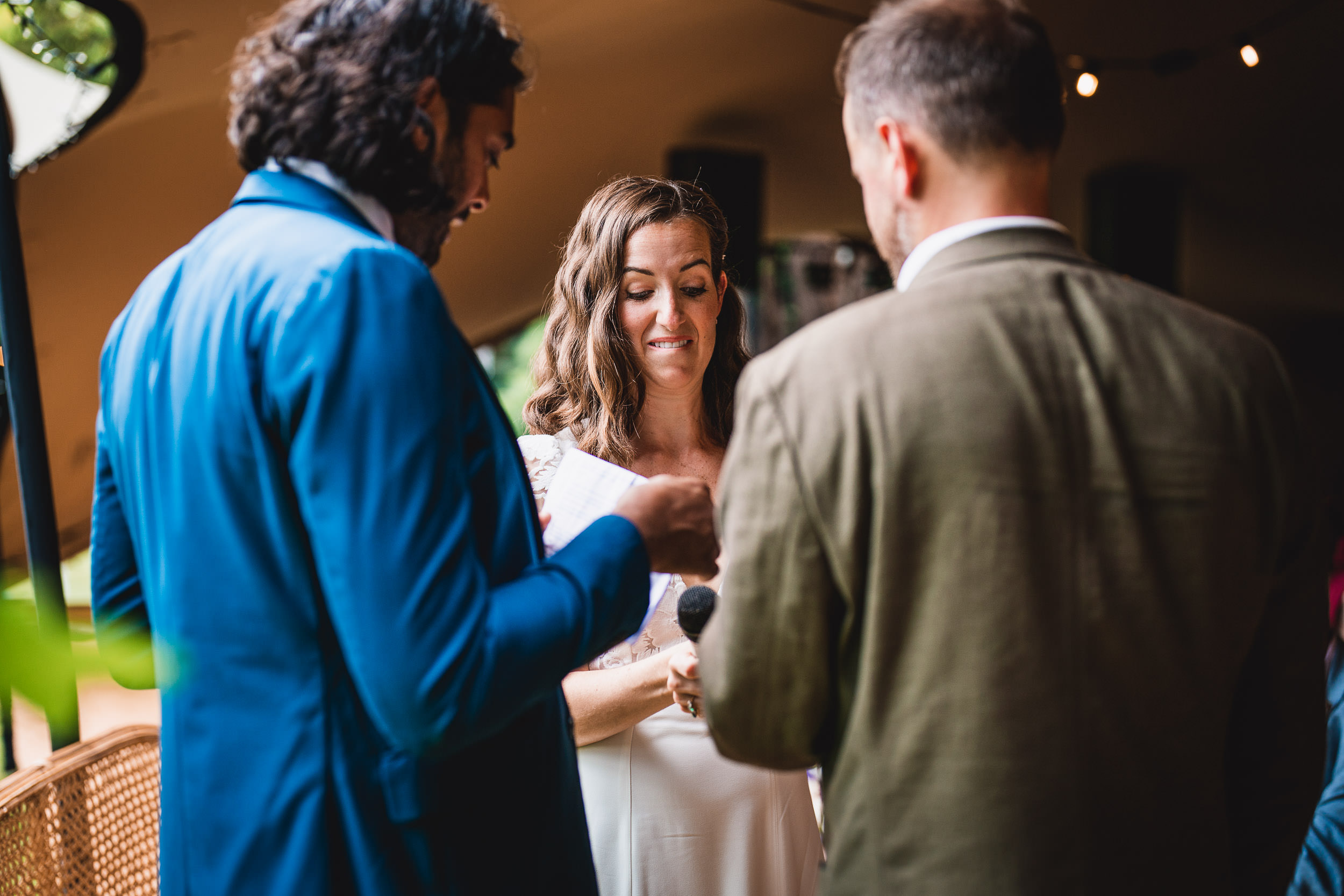 Two men and a woman in formal attire are standing together at a wedding ceremony. The woman holds a microphone.