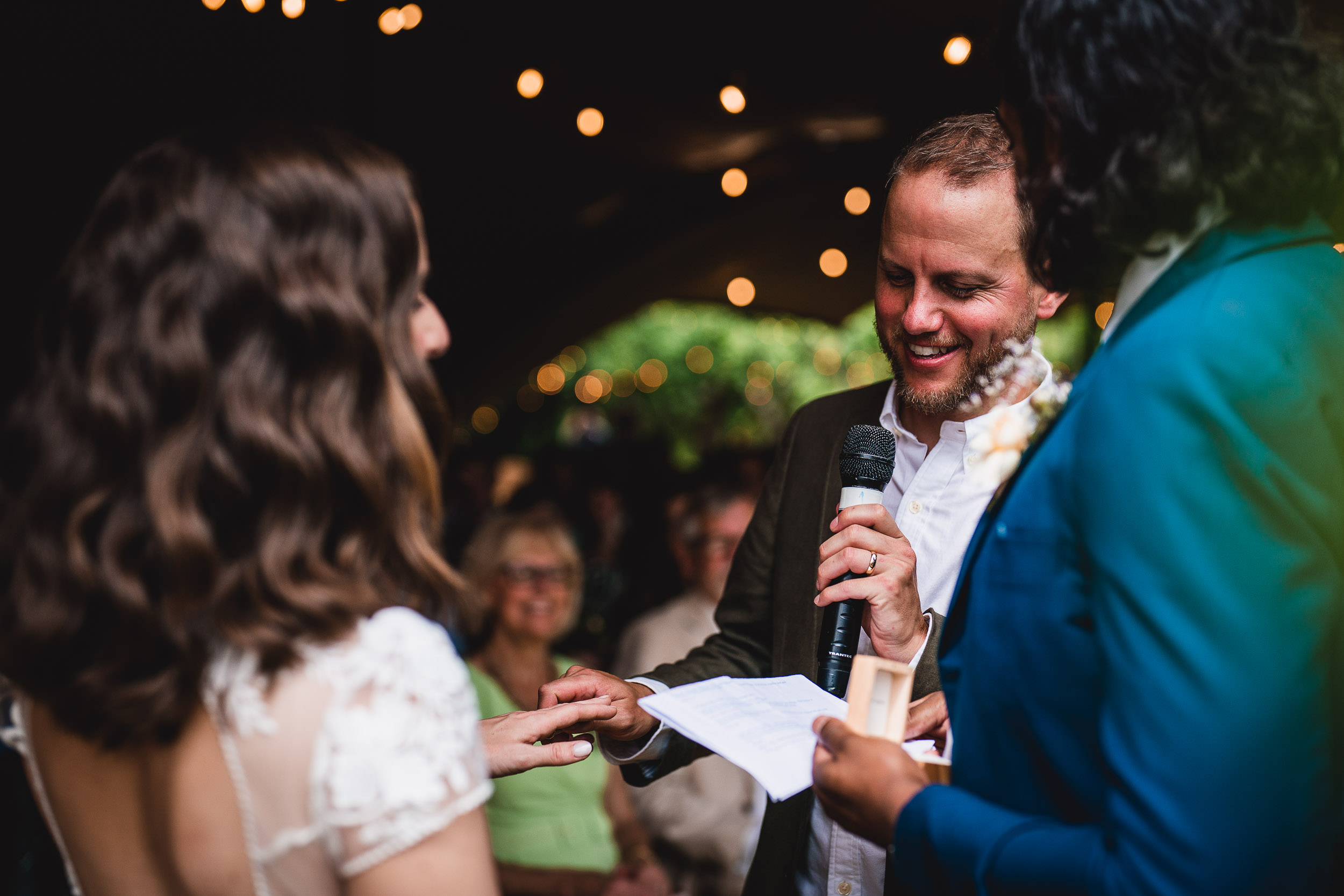 A man and woman exchange rings in a ceremony. Another person in a blue suit officiates, holding a microphone and paper. String lights illuminate the background.