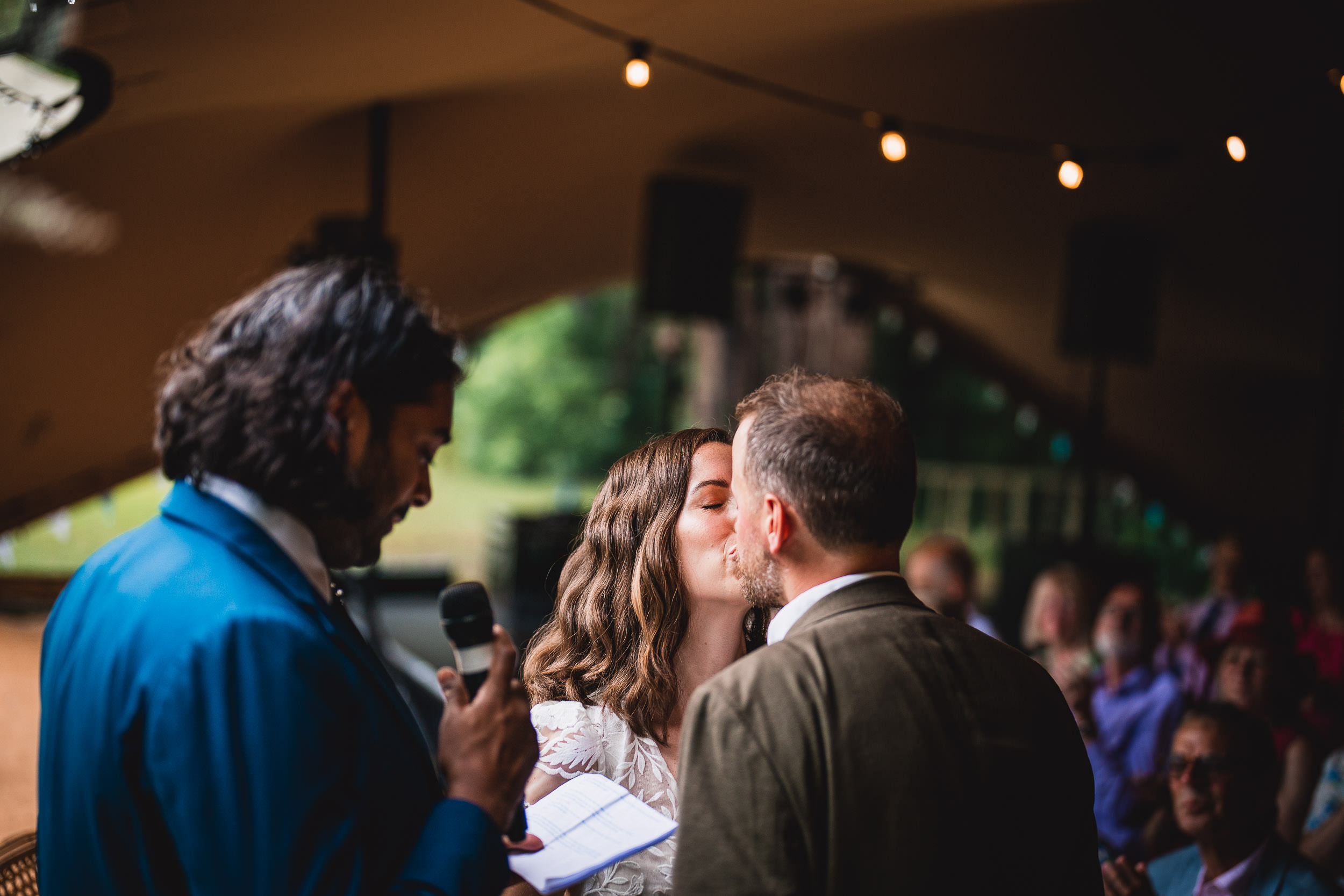 A couple shares a kiss during their wedding ceremony as an officiant holds a microphone and speaks. Guests are seated in the background under string lights.