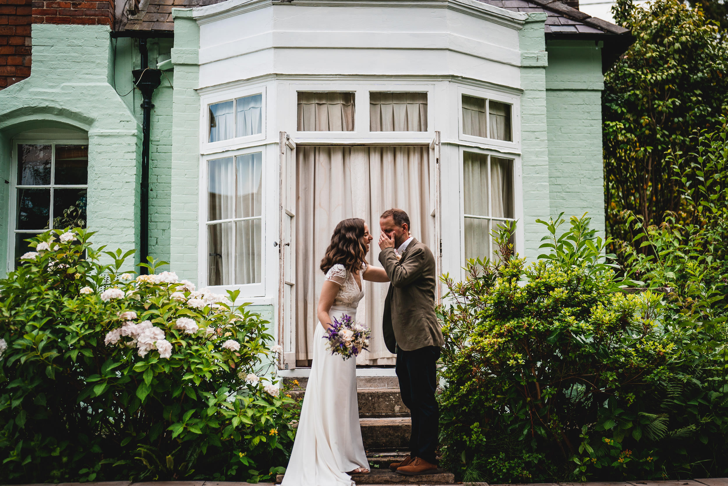 A couple stands facing each other outside a house with green walls and a bay window. The woman holds a bouquet, and both are surrounded by lush greenery.