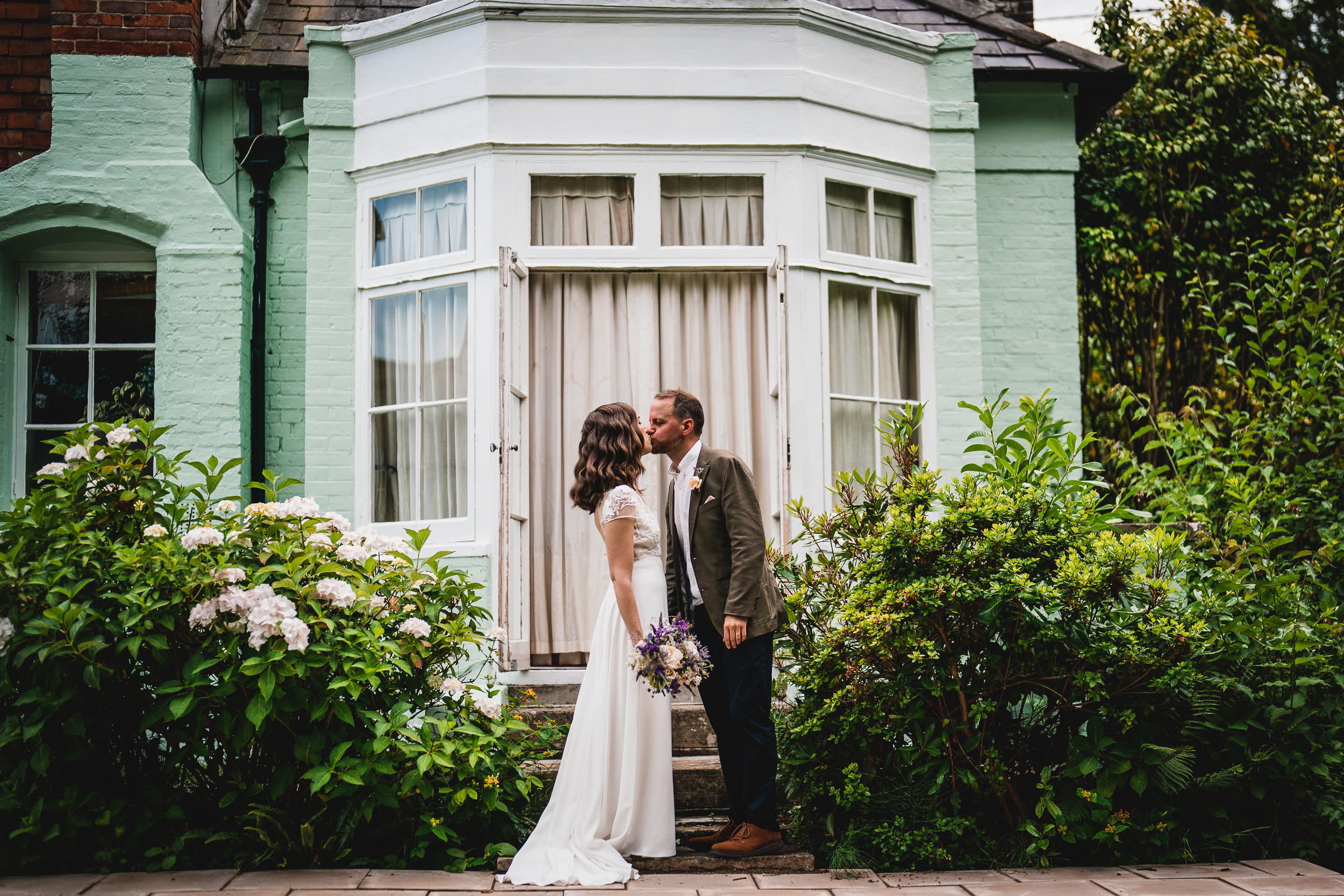 A couple kisses in front of a green house with a bay window, surrounded by lush plants and flowers. The woman holds a bouquet.