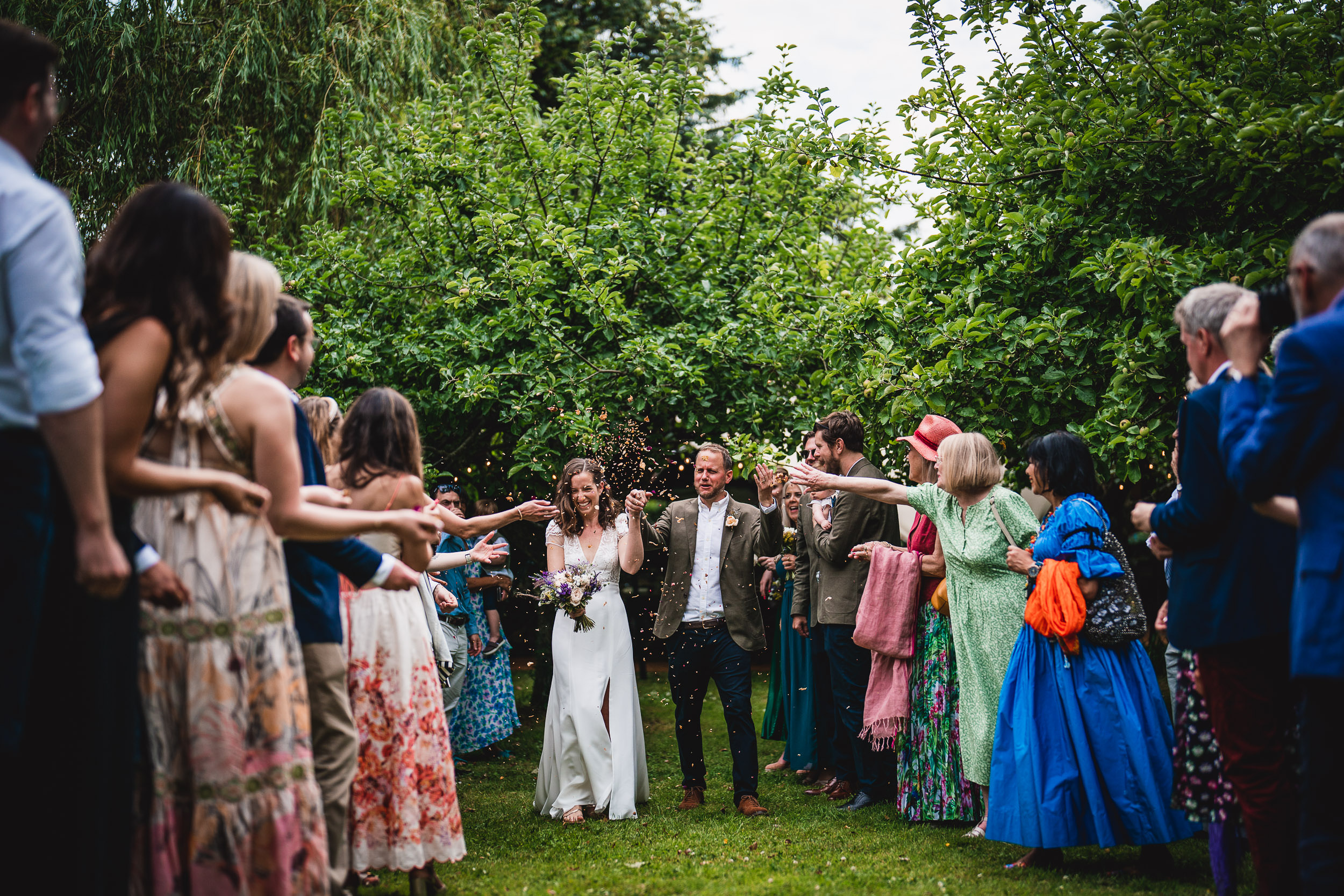 A bride and groom walk through a crowd of people outdoors. Guests on both sides throw confetti as the couple smiles and holds hands. Greenery surrounds the scene.