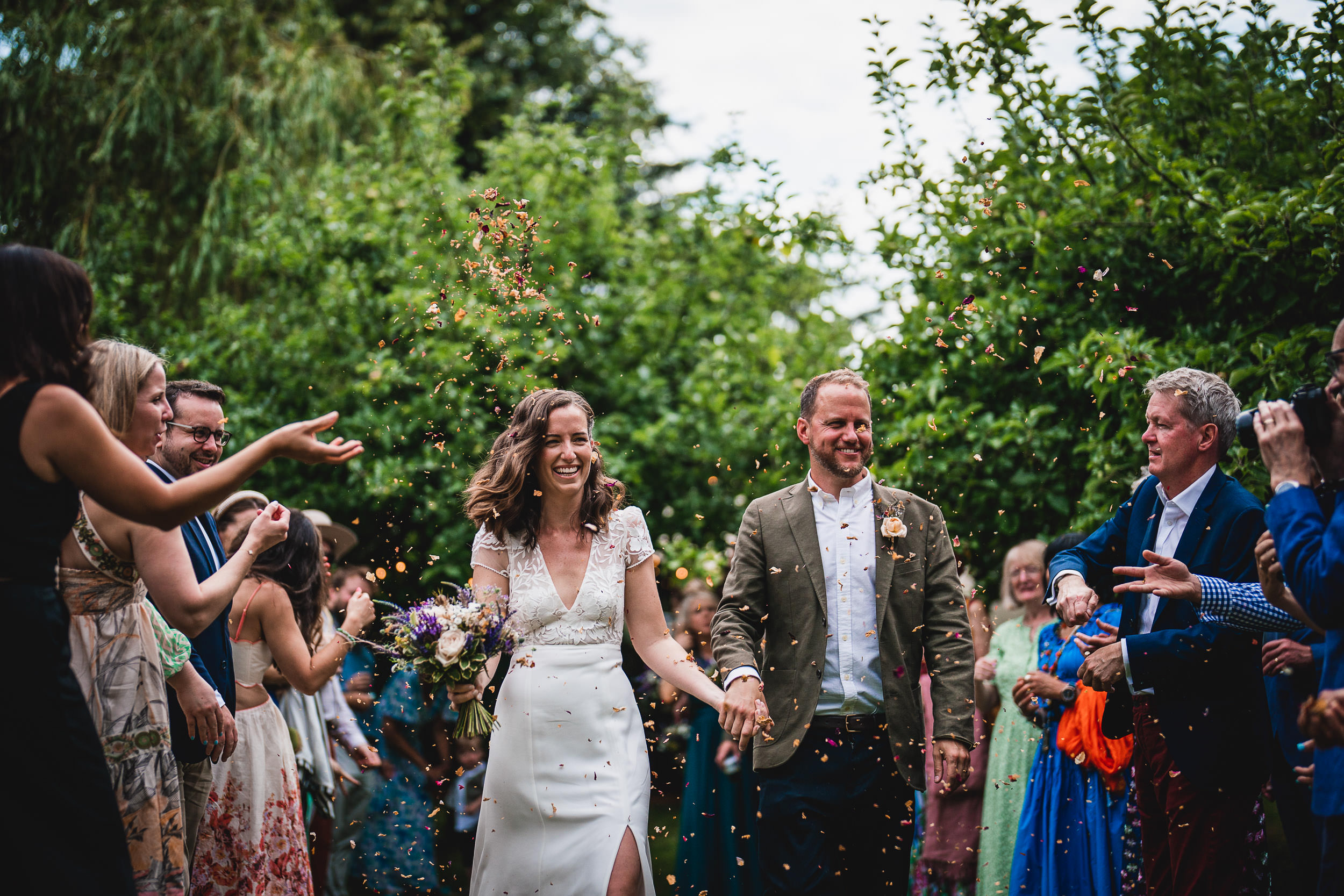 A bride and groom walk down an outdoor aisle while guests celebrate and throw confetti. The bride holds a bouquet and smiles.