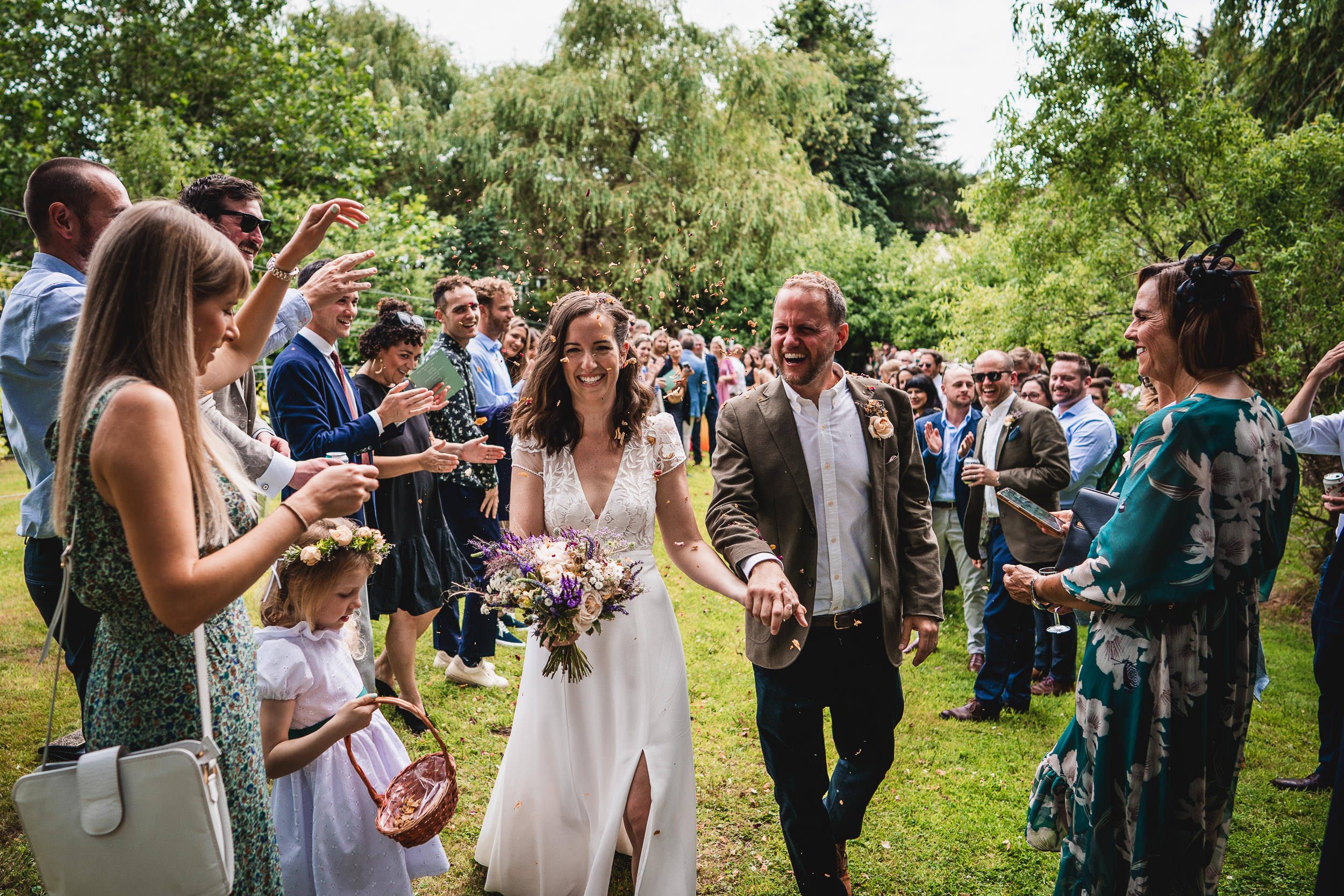 A bride and groom walk down a grassy aisle surrounded by cheering guests in a garden setting. The bride holds a bouquet and the groom wears a jacket; children are present.