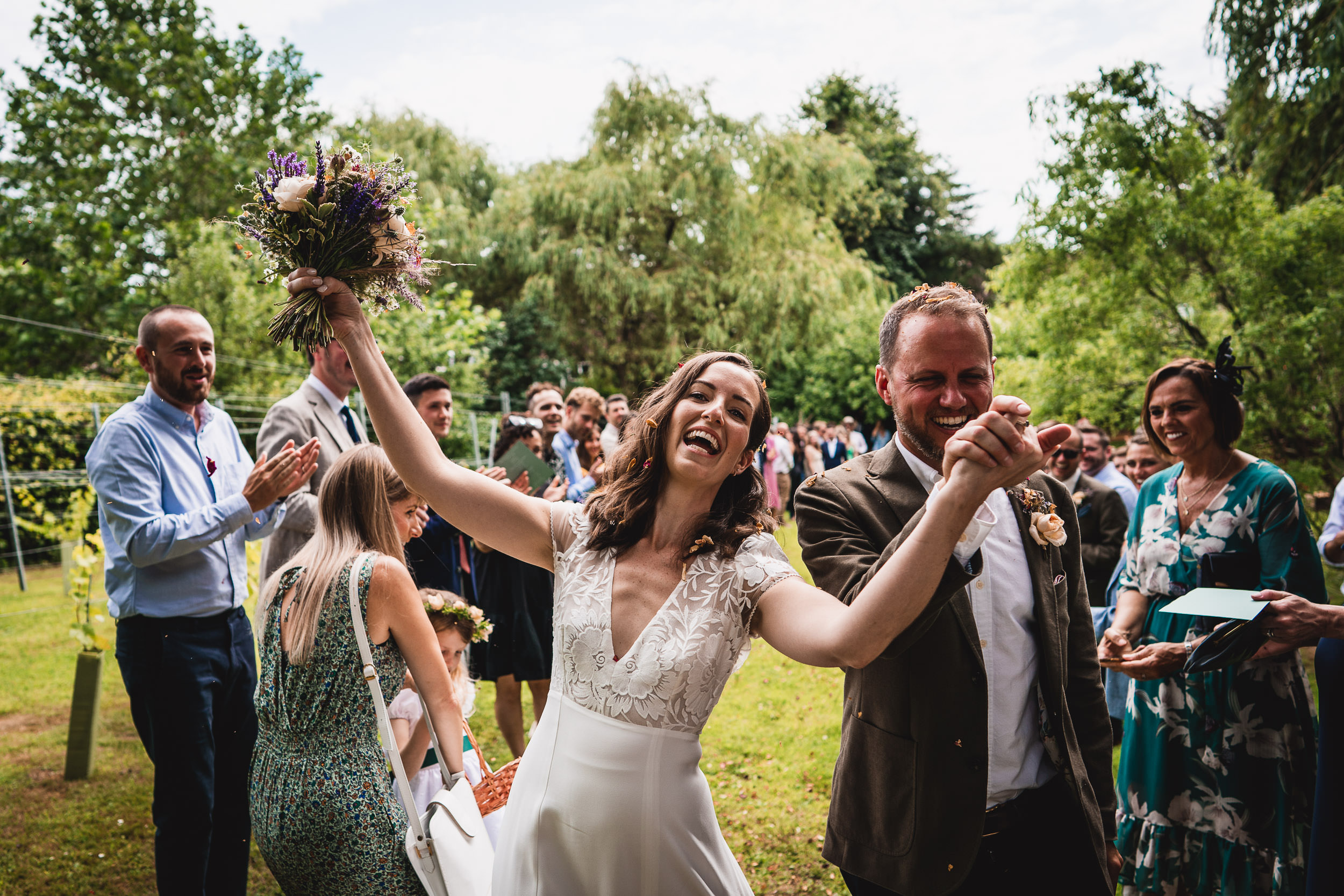 A joyful bride and groom celebrate outdoors with guests applauding in the background, surrounded by lush greenery.