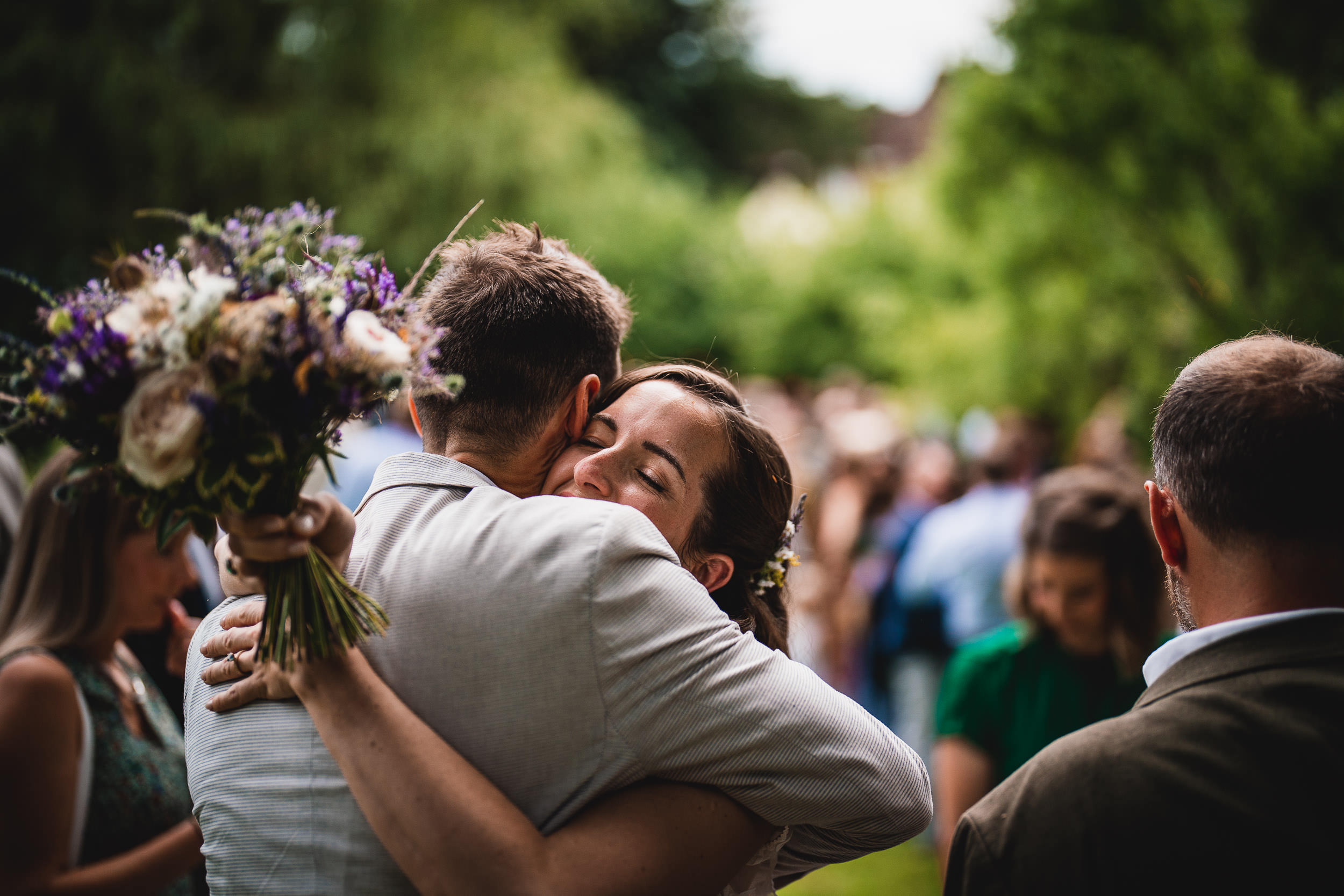 A couple embraces outdoors, surrounded by people. One holds a bouquet of flowers.