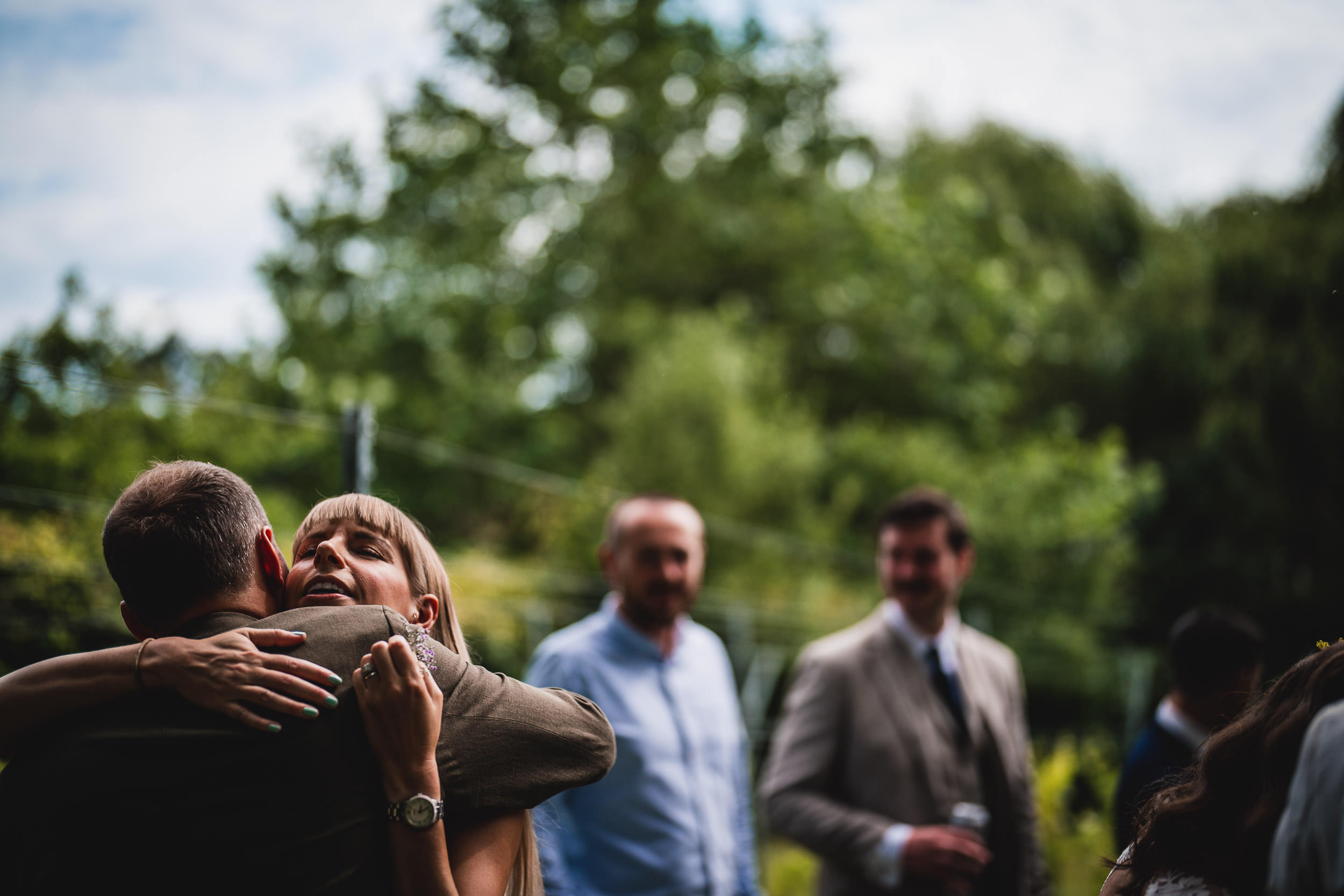 A woman and a man are embracing outdoors, with a blurred background of trees and people.