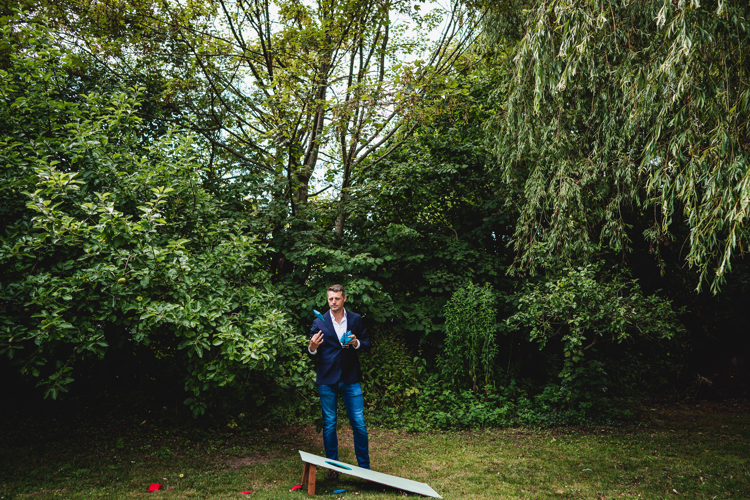 A man in a suit stands in a lush green garden near a wooden board, gesturing with his hands.