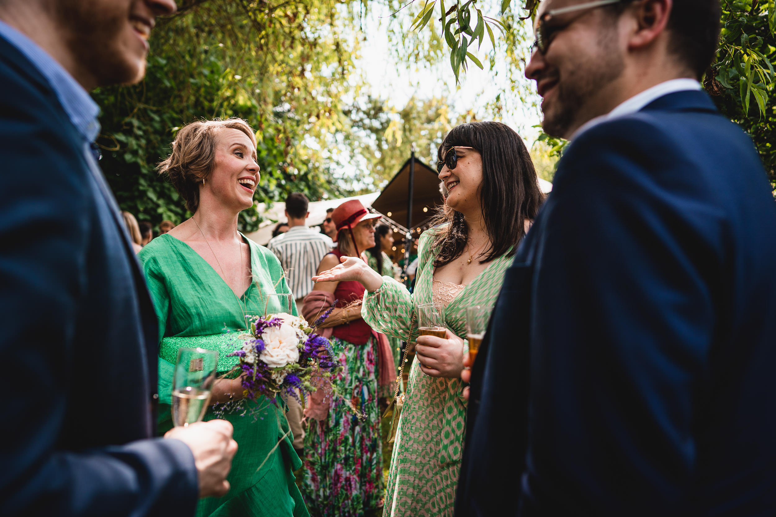 A group of people in formal attire enjoying a conversation outdoors, with two women holding drinks and one holding a small bouquet.