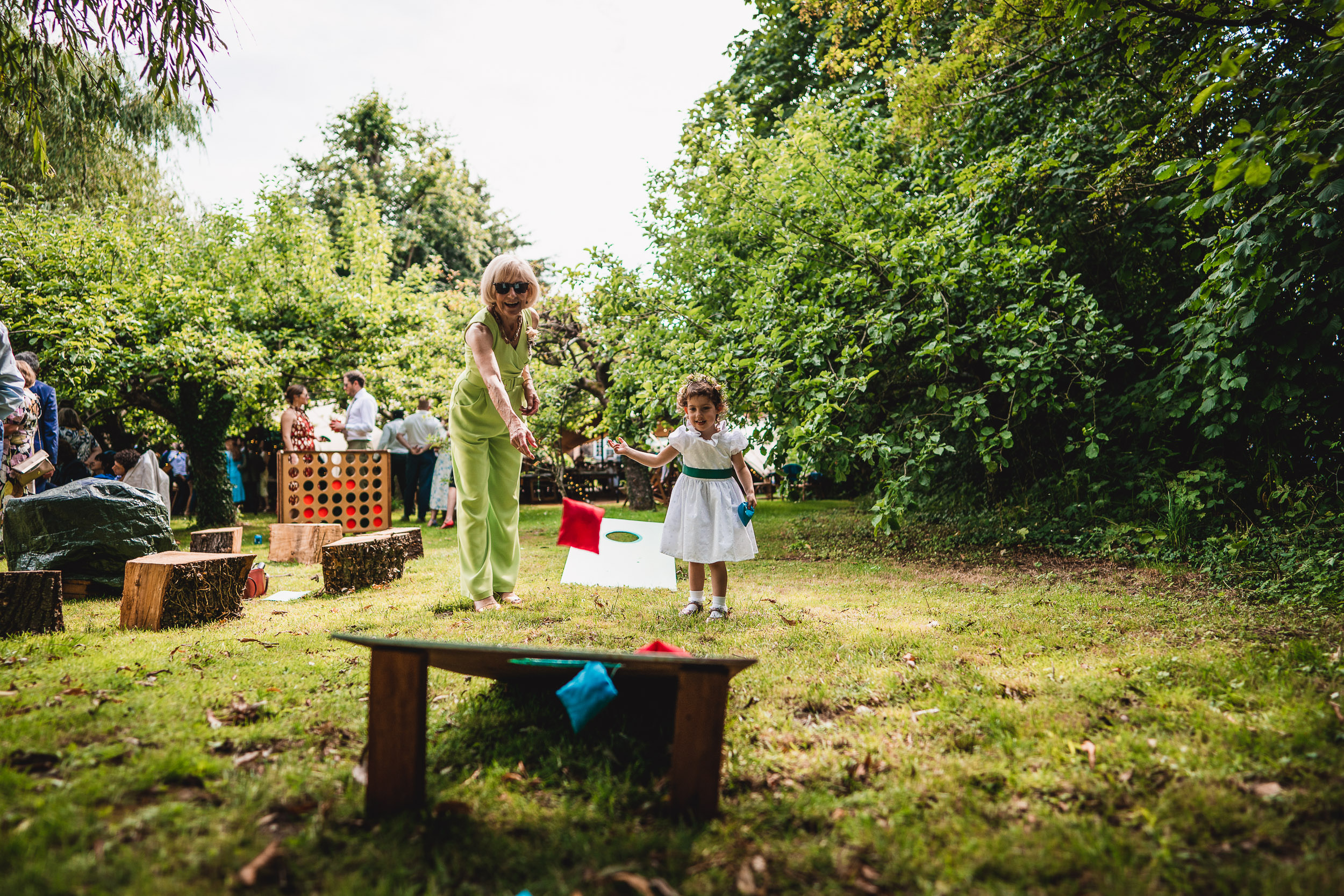 An adult and child throw bean bags outdoors in a garden. People and trees in the background.