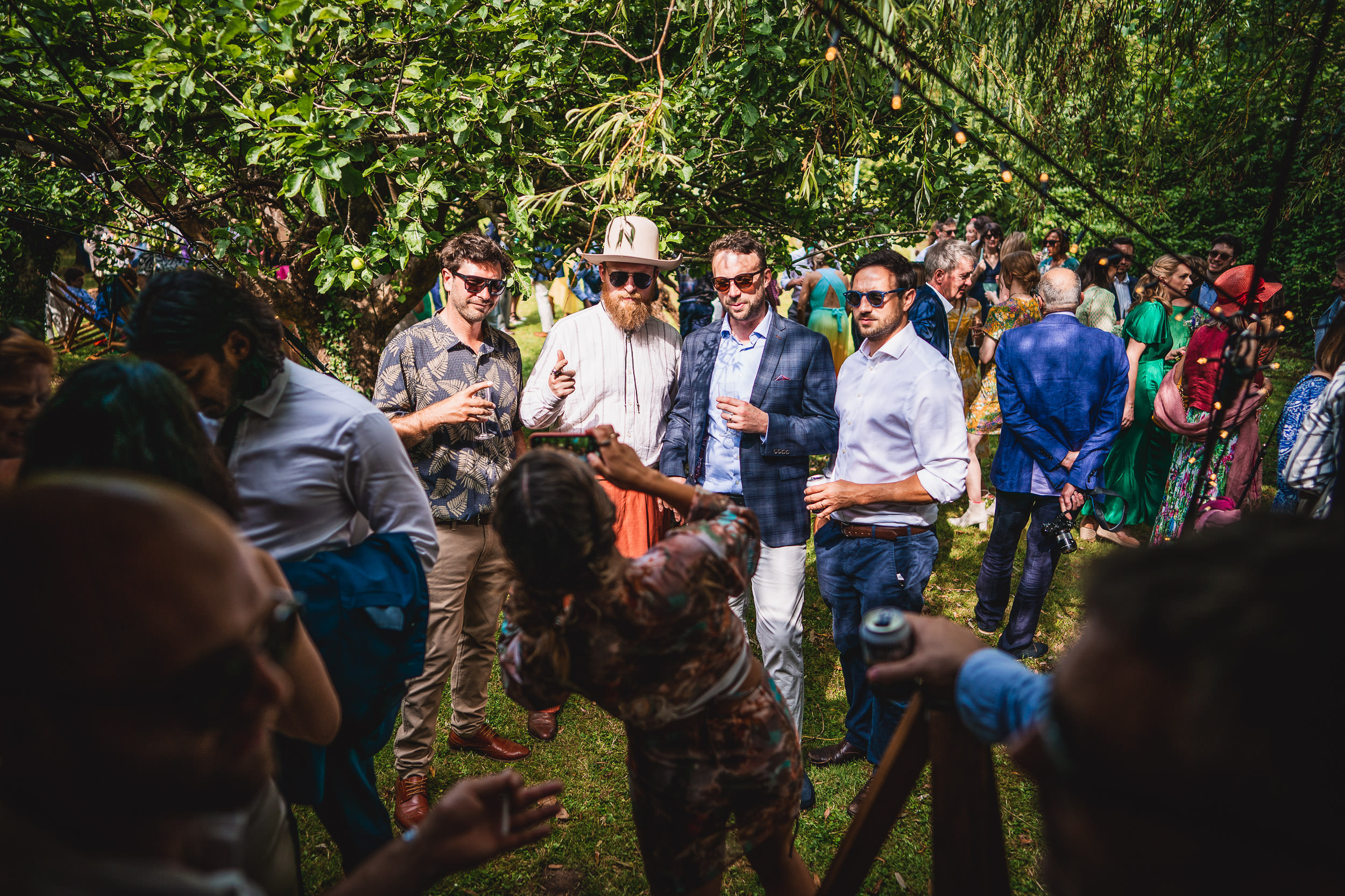 Group of people gather outdoors in a garden setting, with some posing for a photo. They are casually dressed, and the scene is lively with greenery around.