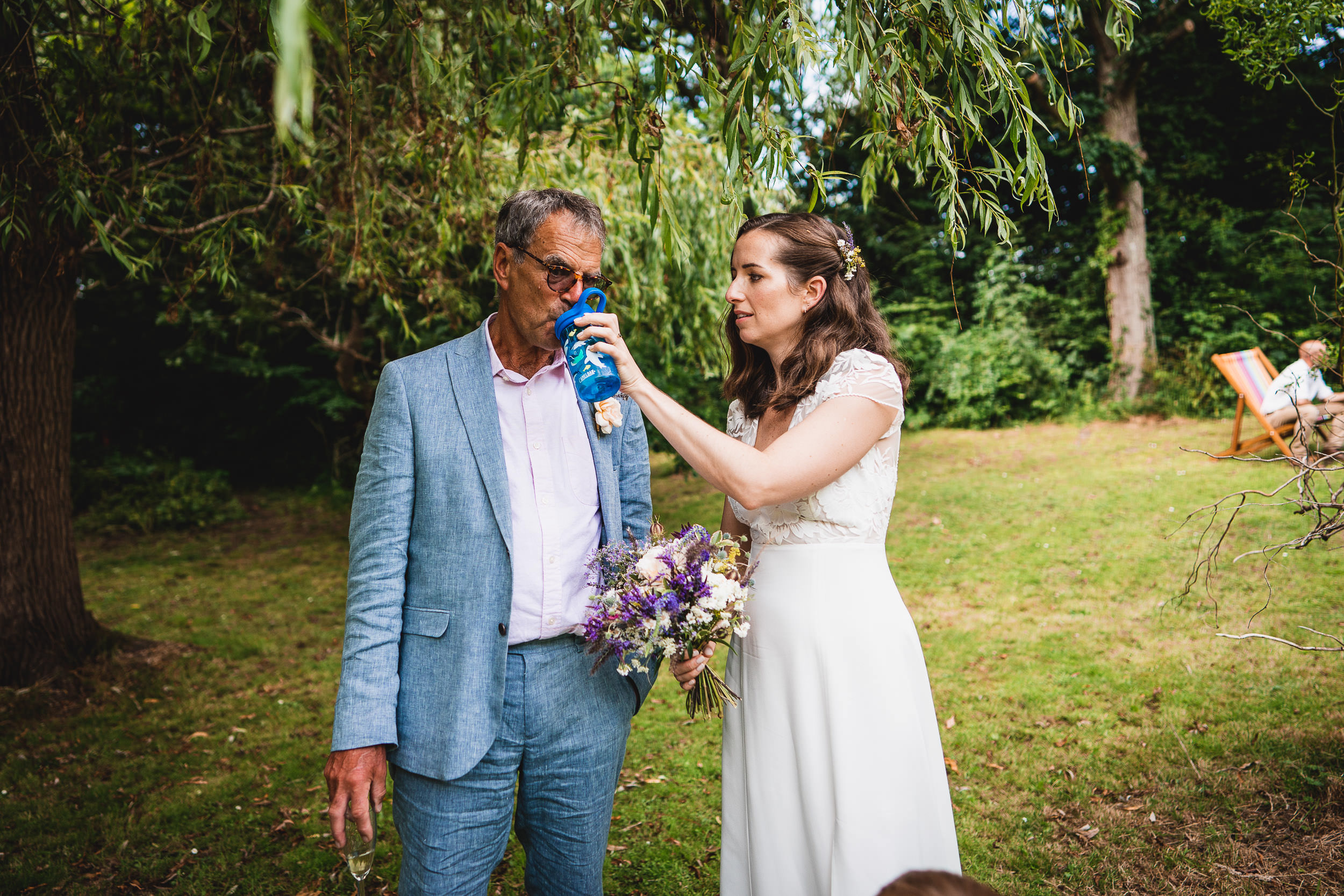 A woman in a white dress sprays water on a man's face outdoors.