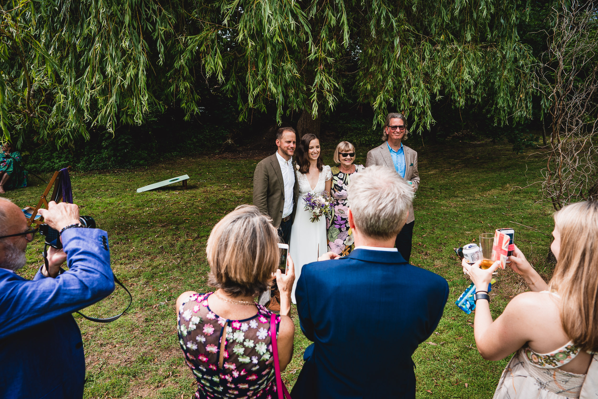 A group of people taking photos at an outdoor wedding under a large tree, with a bride and groom smiling in the center.