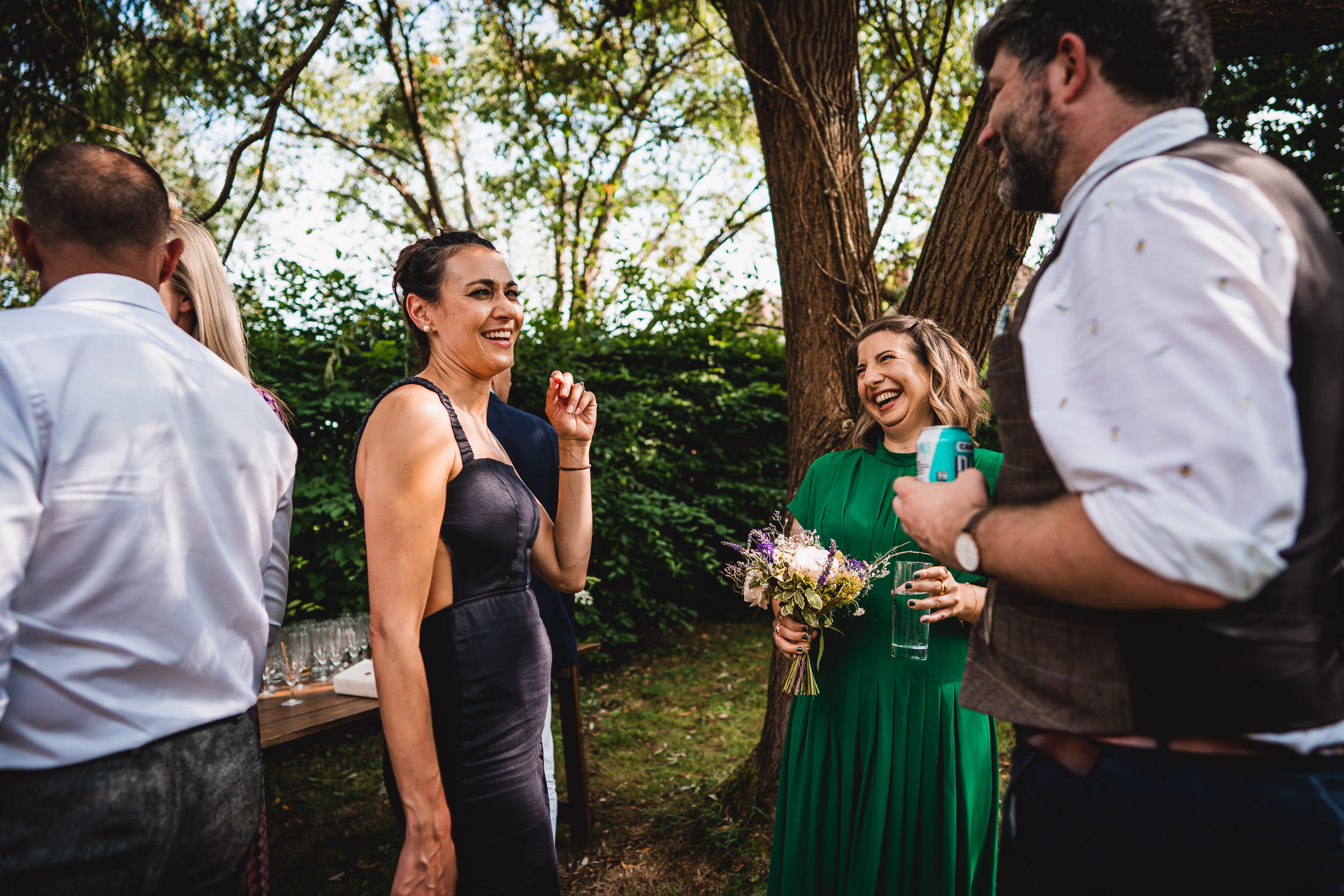 A group of people outdoors, smiling and talking. One holds a bouquet, another a drink. Trees and greenery are in the background.