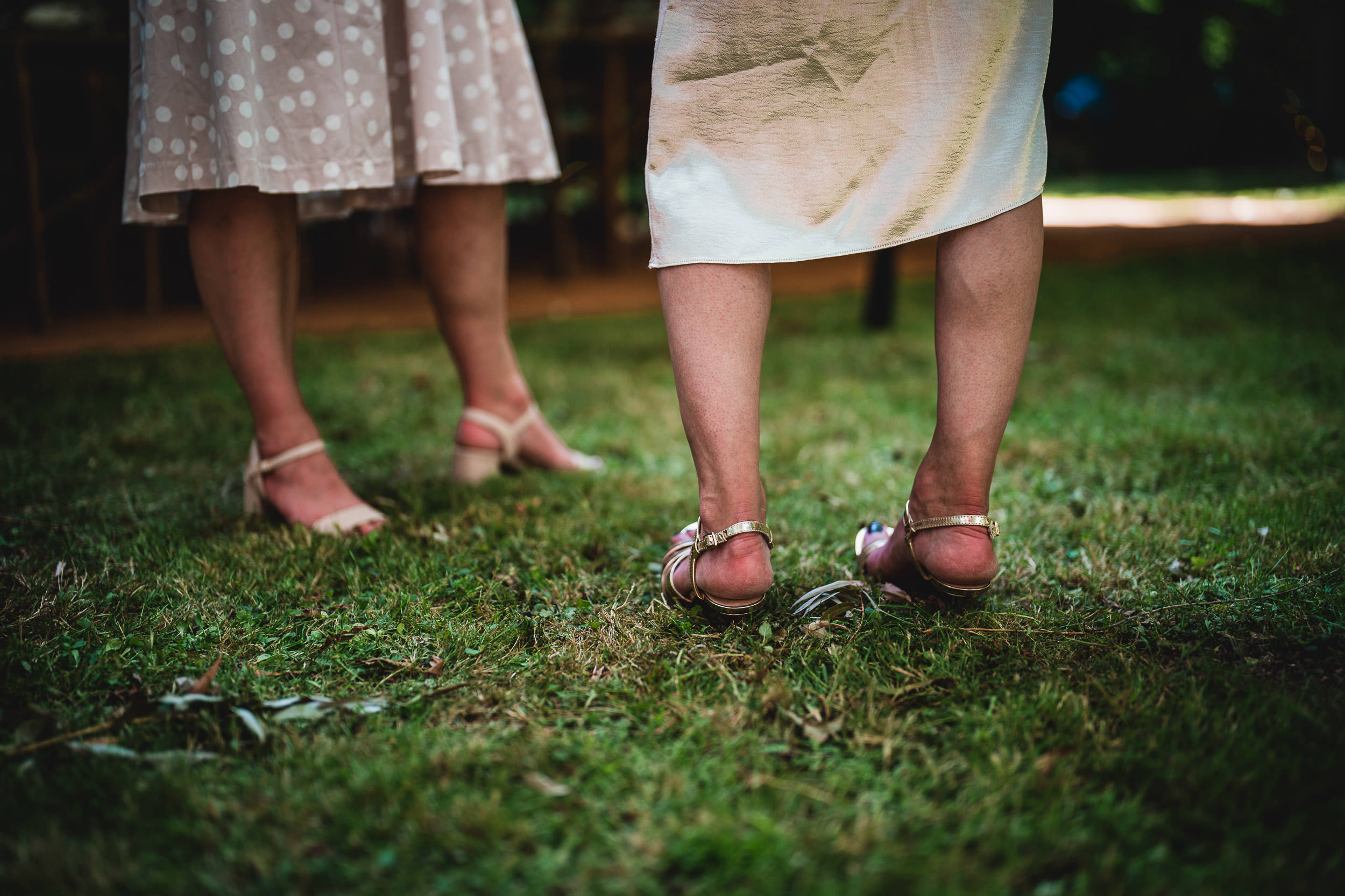 Two people in dresses and sandals stand on a grassy area.