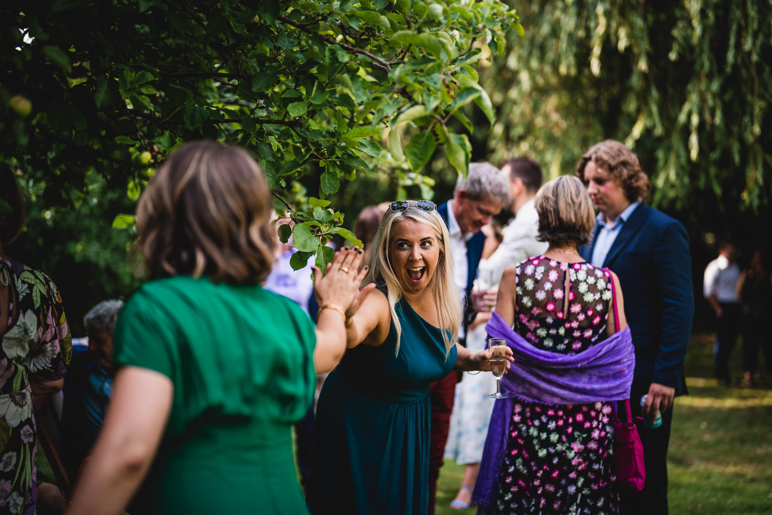 A woman in a blue dress high-fives another woman in a green dress at an outdoor gathering. People in the background are socializing.