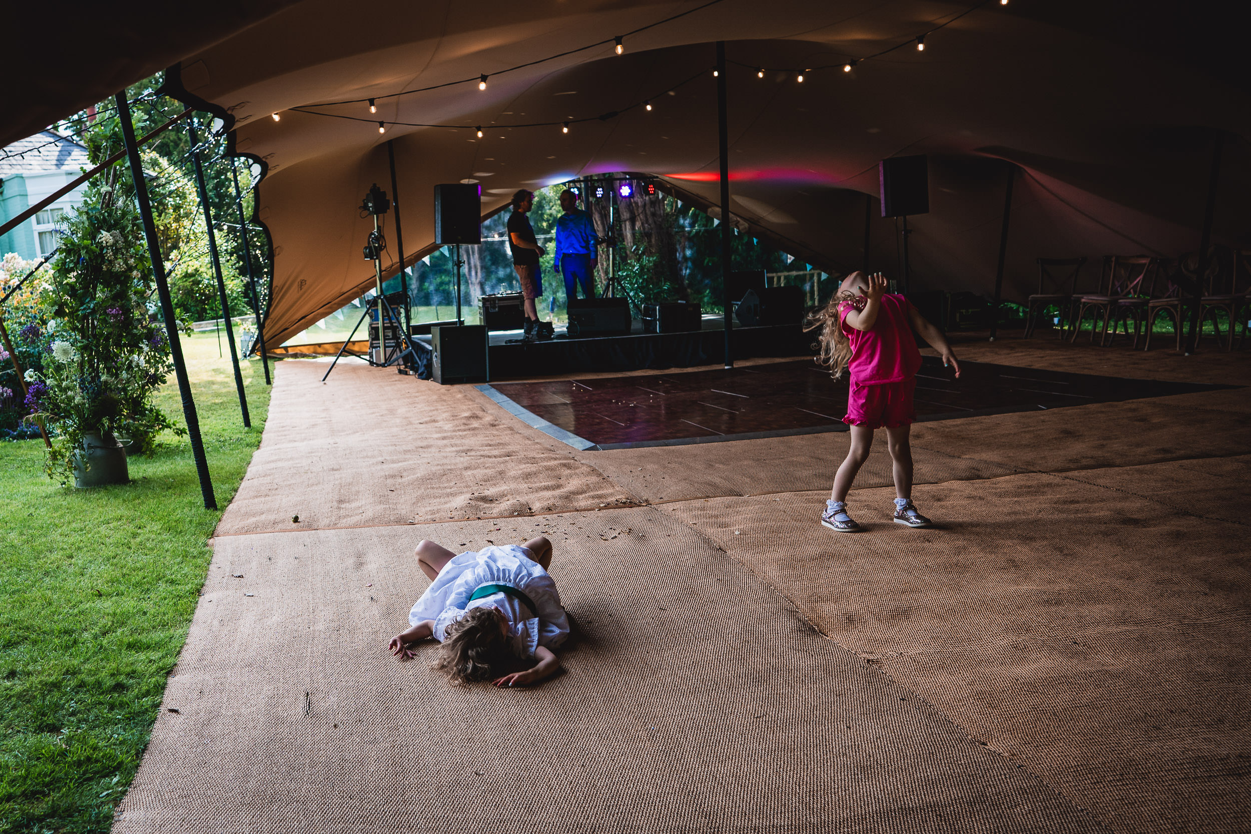 Two children play under a large tent with a wooden stage, one lying on the ground and the other standing, with string lights above.