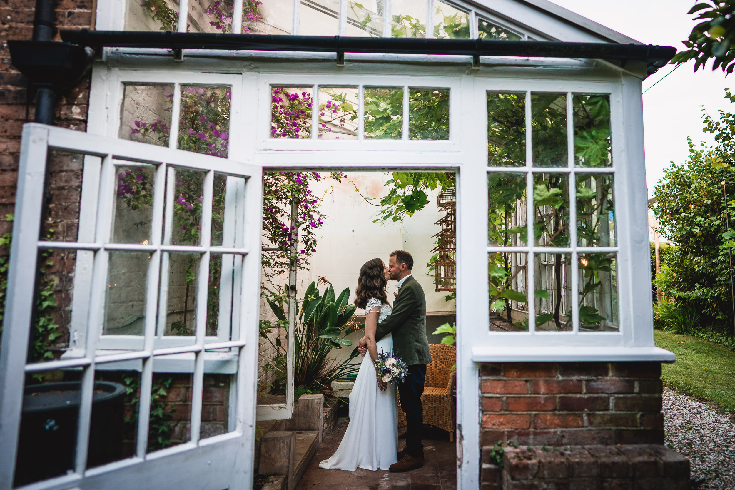 A couple embraces inside a glasshouse adorned with greenery and purple flowers. The bride holds a bouquet, and the open door offers a view of the garden outside.