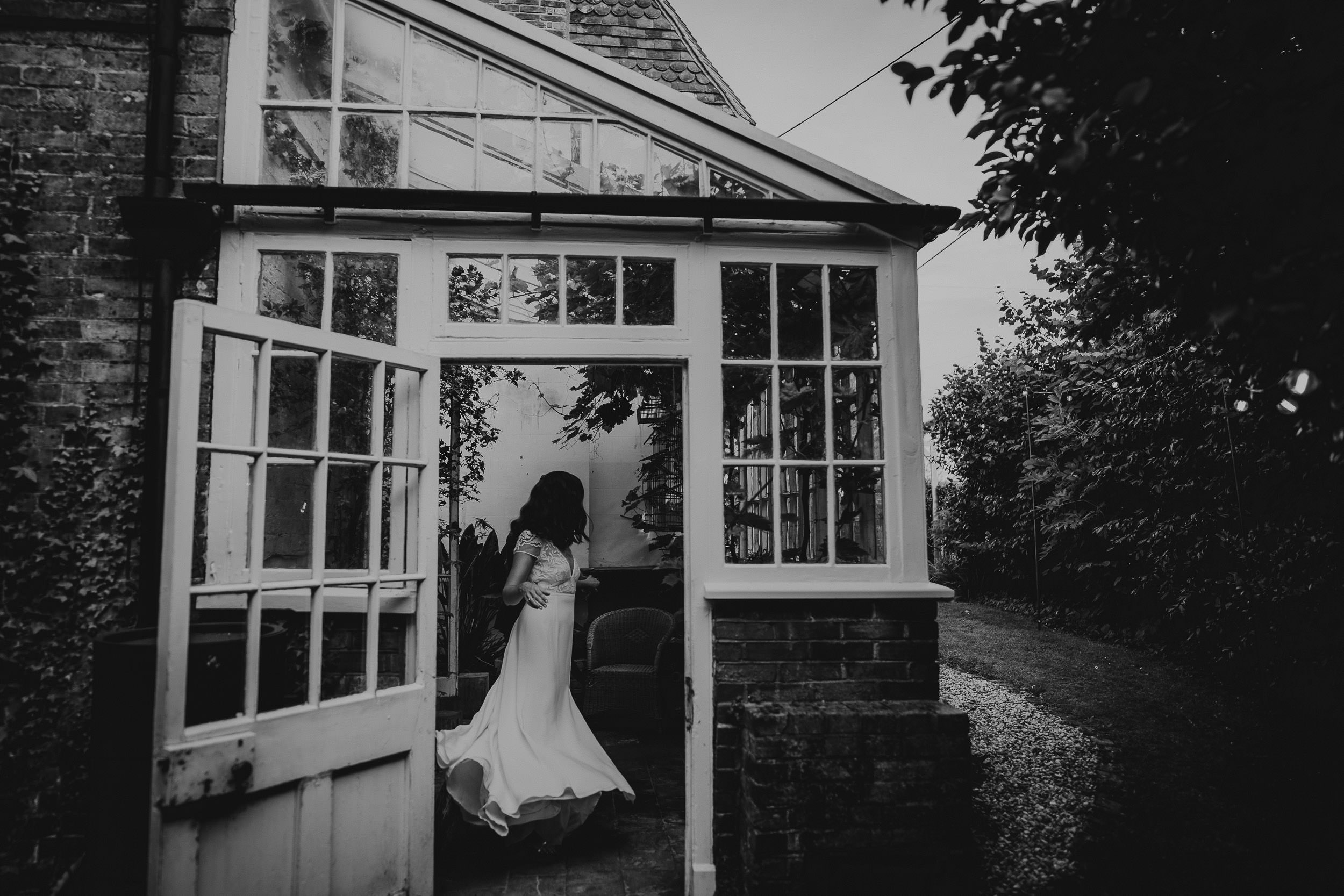 A person in a flowing dress enters a brick and glass building with an open door. The scene is surrounded by trees and foliage.