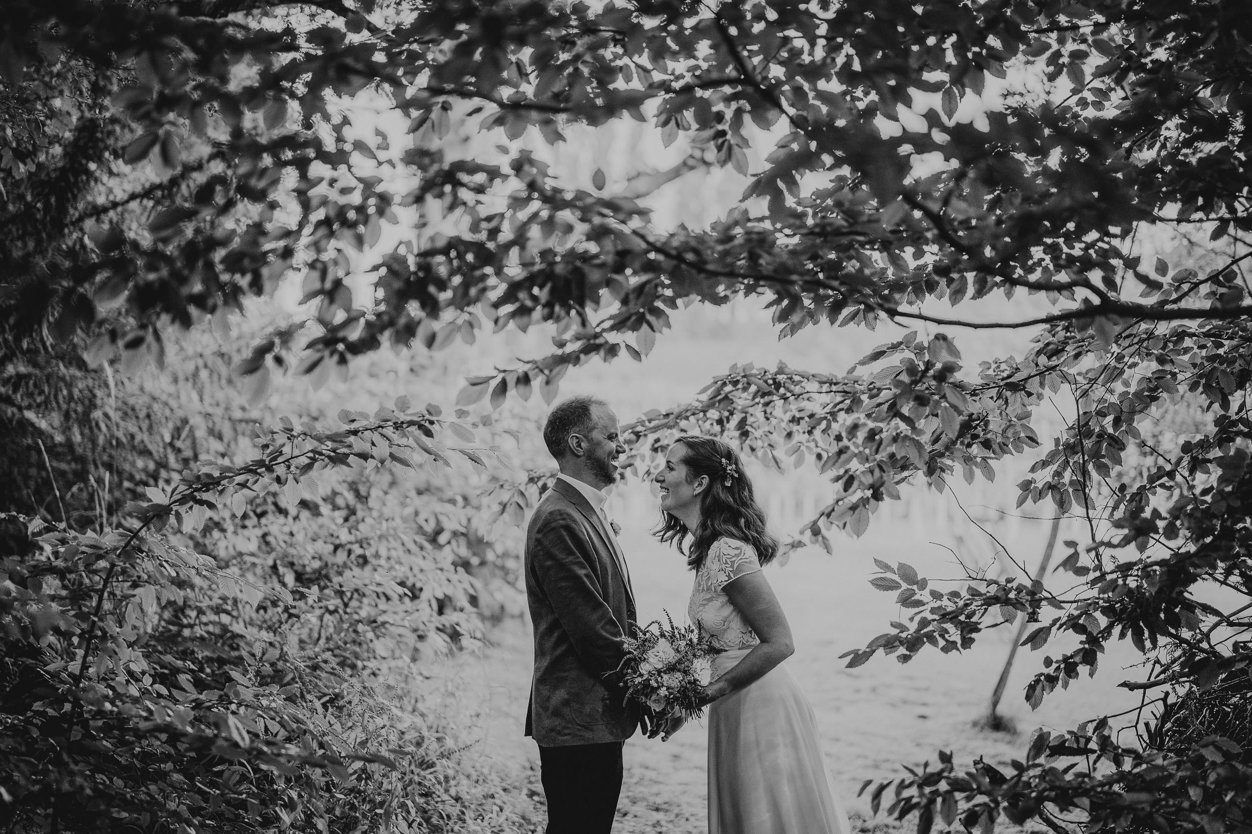 A couple stands under tree branches, holding hands, in a black-and-white outdoor setting.