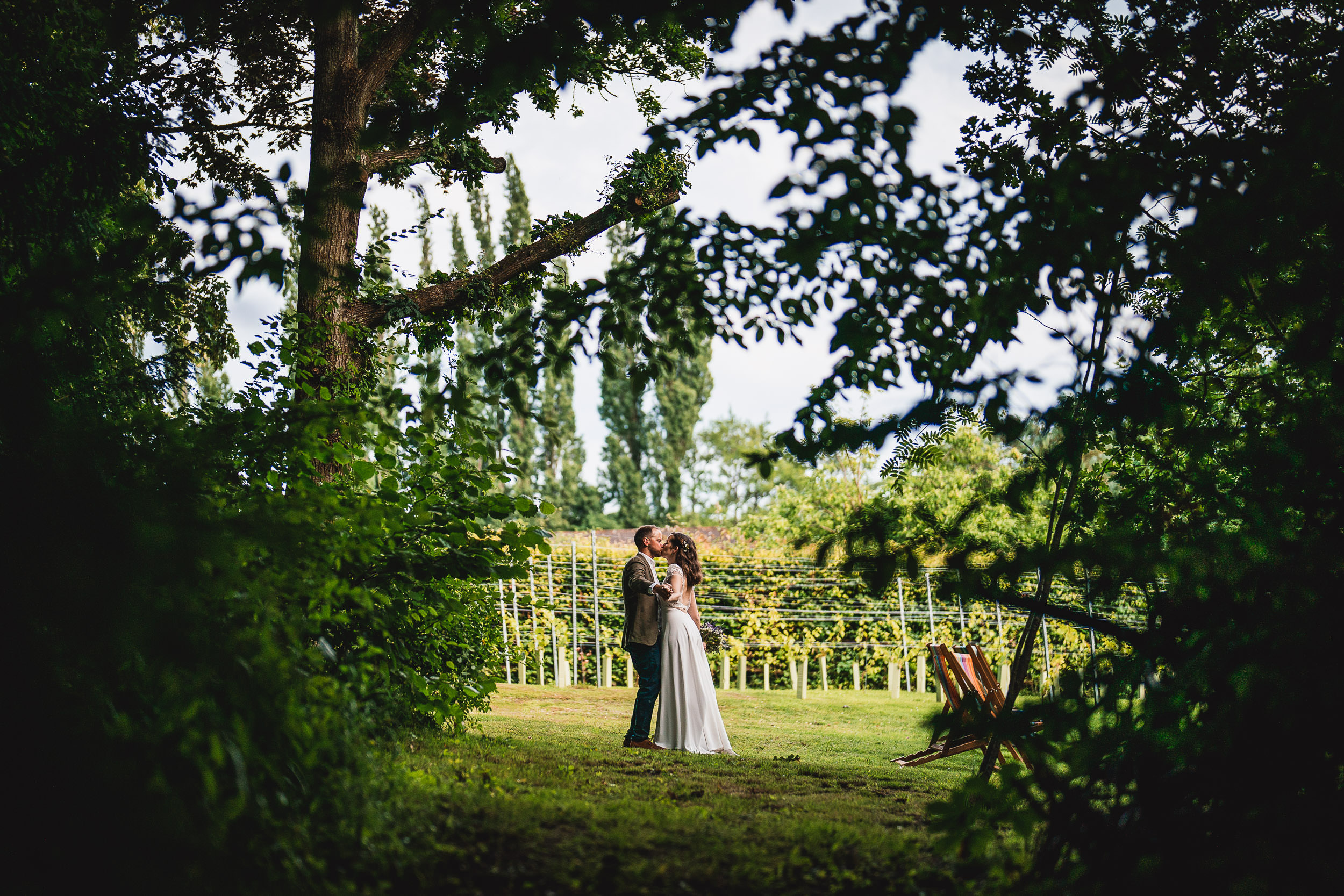 A couple in wedding attire kisses in a lush, green outdoor setting surrounded by trees and a vineyard in the background.