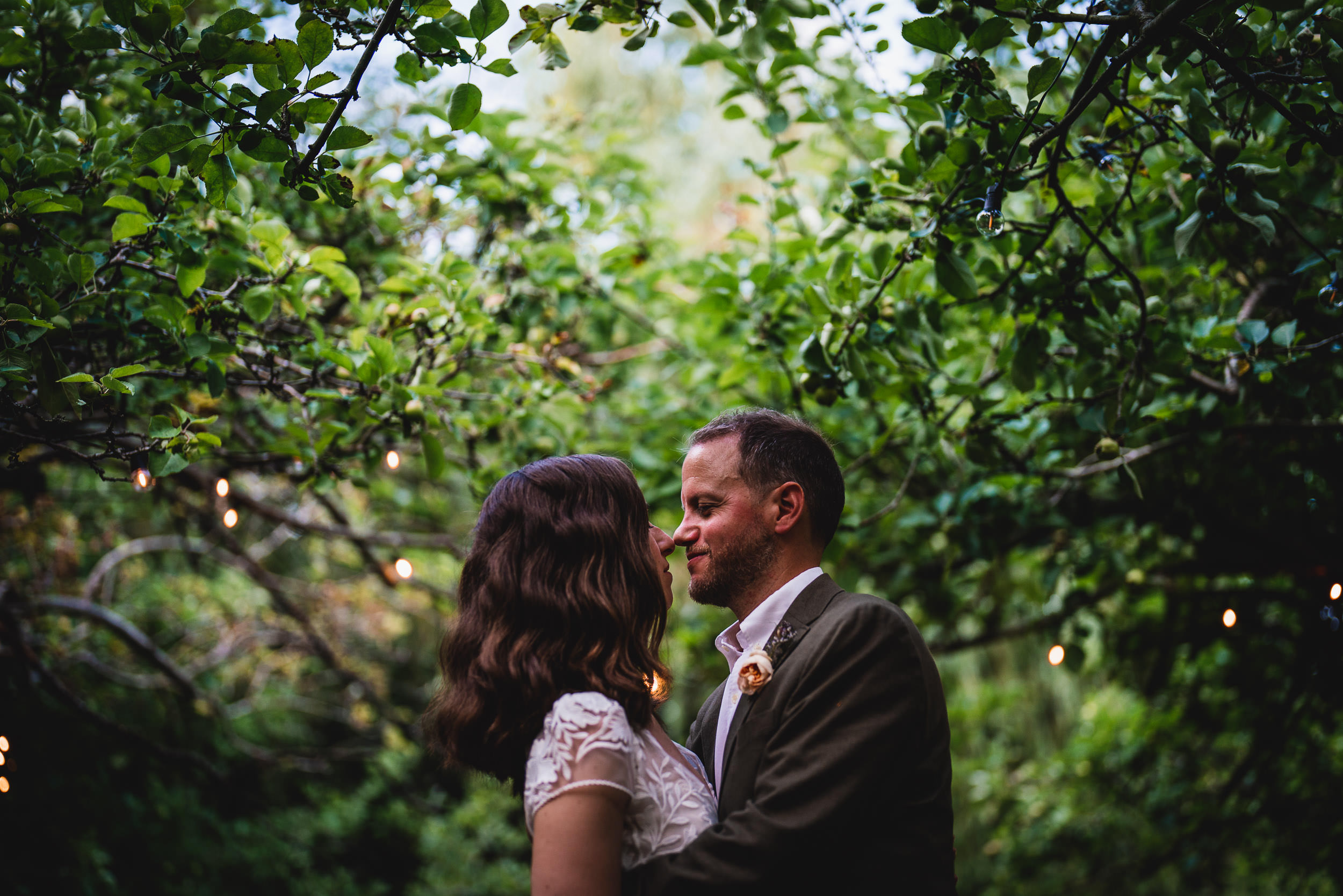 A couple embraces under a canopy of green leaves, with string lights softly glowing in the background.