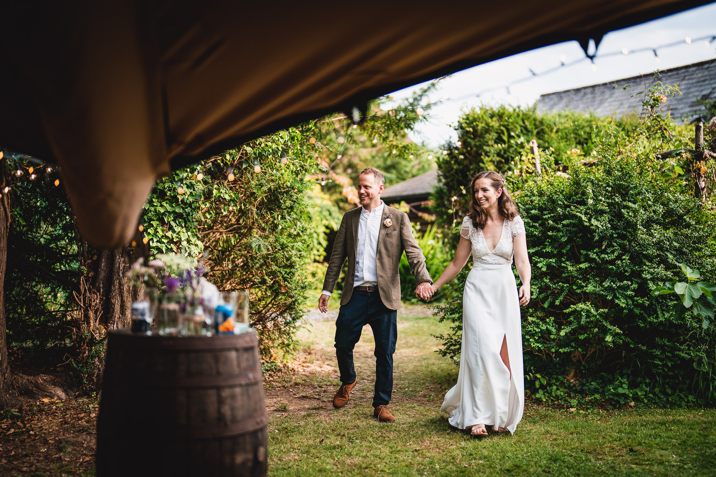 A couple holds hands walking through a garden path, surrounded by greenery and a few decorative lights.