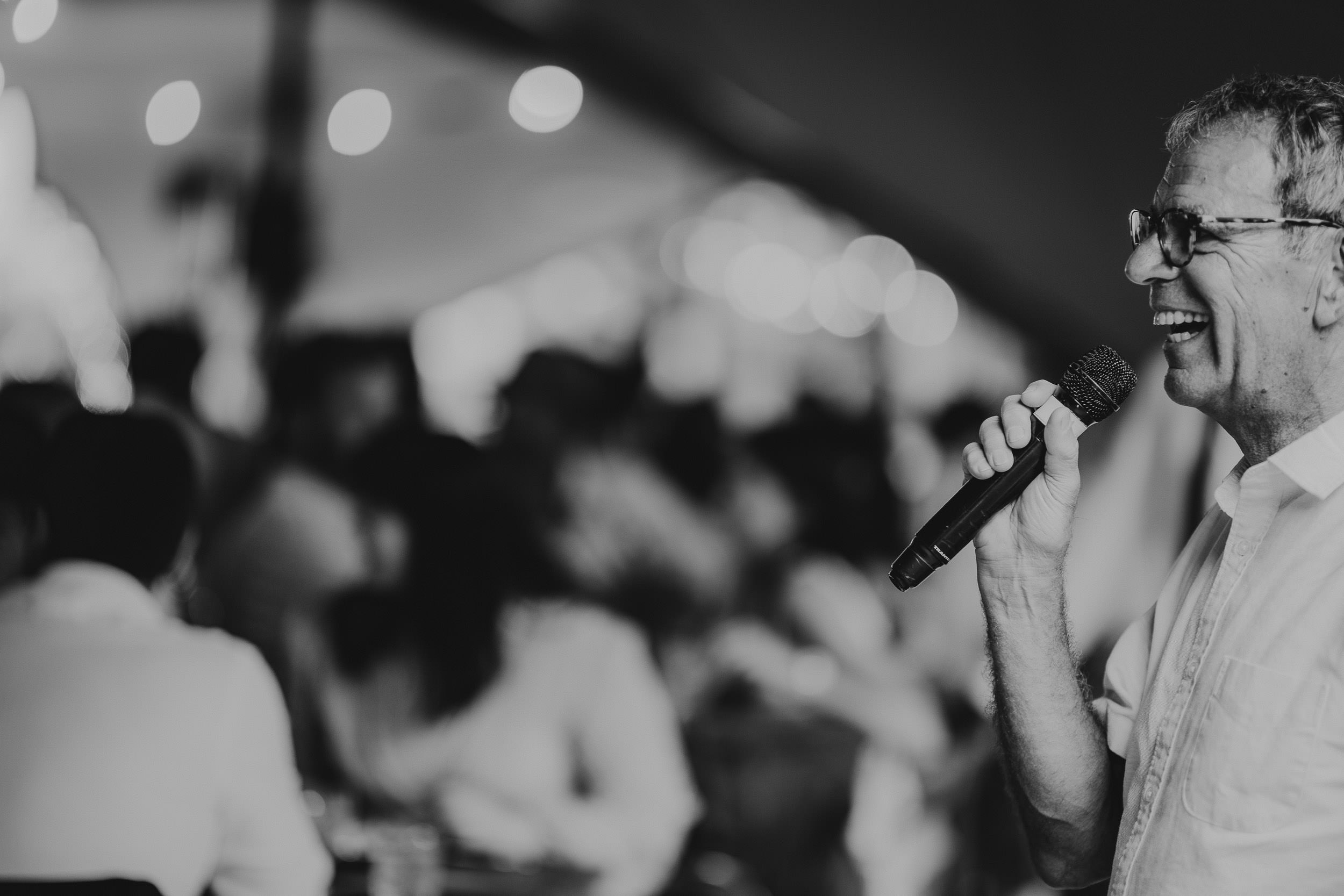 Black and white photo of a person smiling and speaking into a microphone at an indoor event, with a blurred audience in the background.