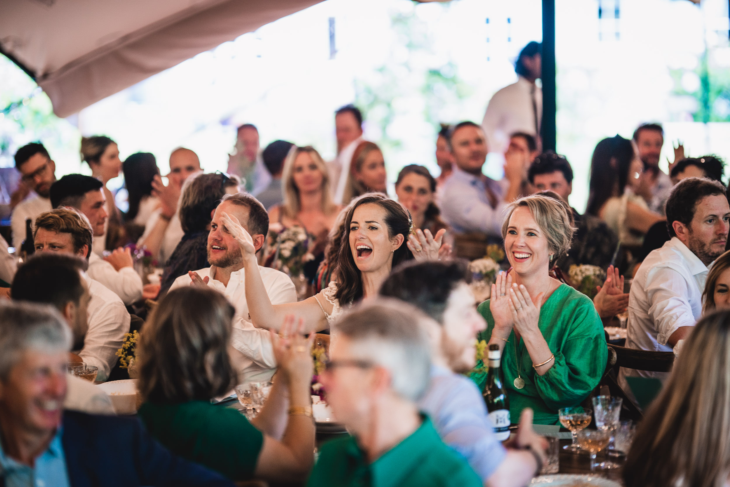 A group of people sit together at an event, clapping and smiling. Tables with drinks and decorations are visible in the background.