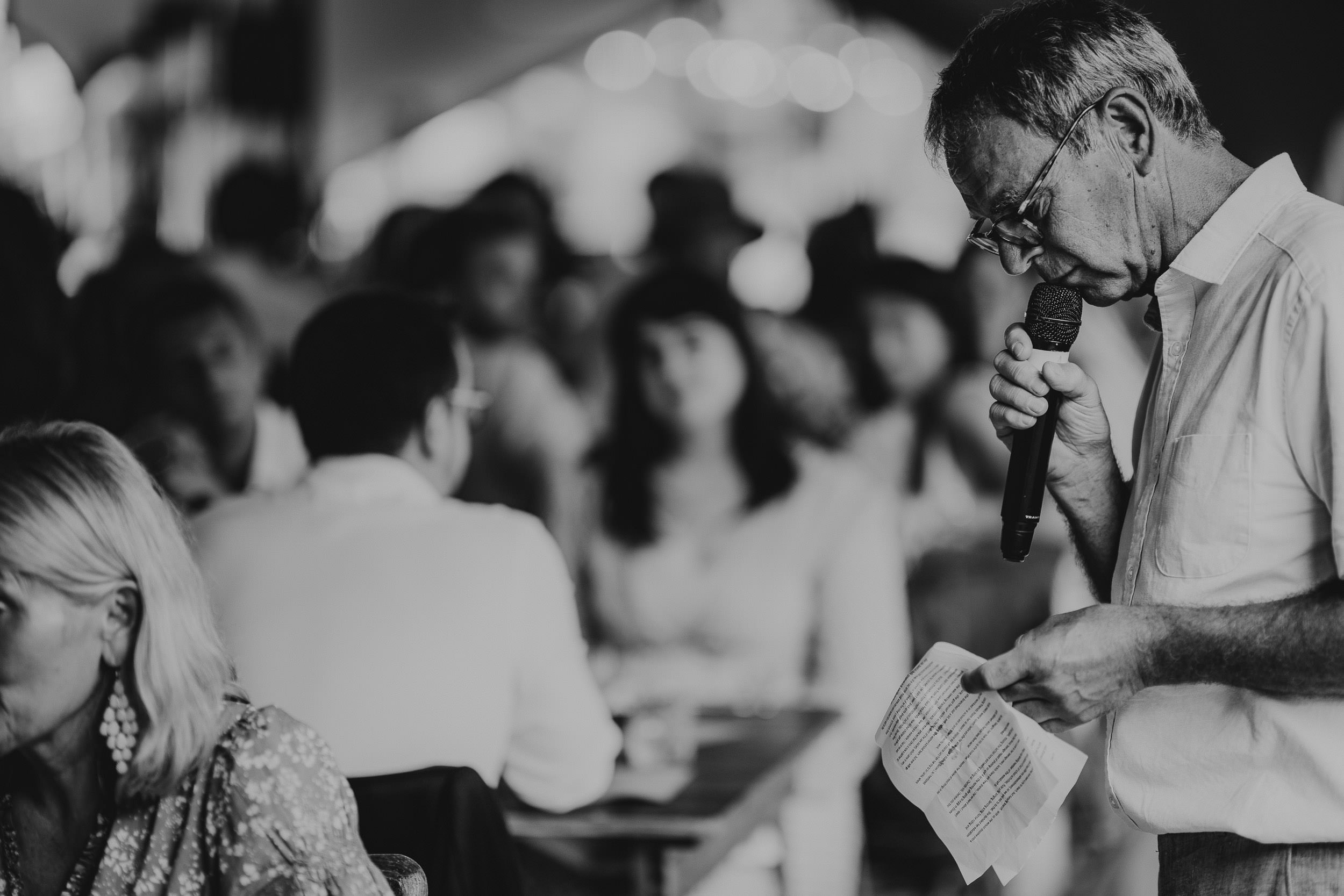 A man in glasses speaks into a microphone while holding a paper, surrounded by a crowd in a blurred background.