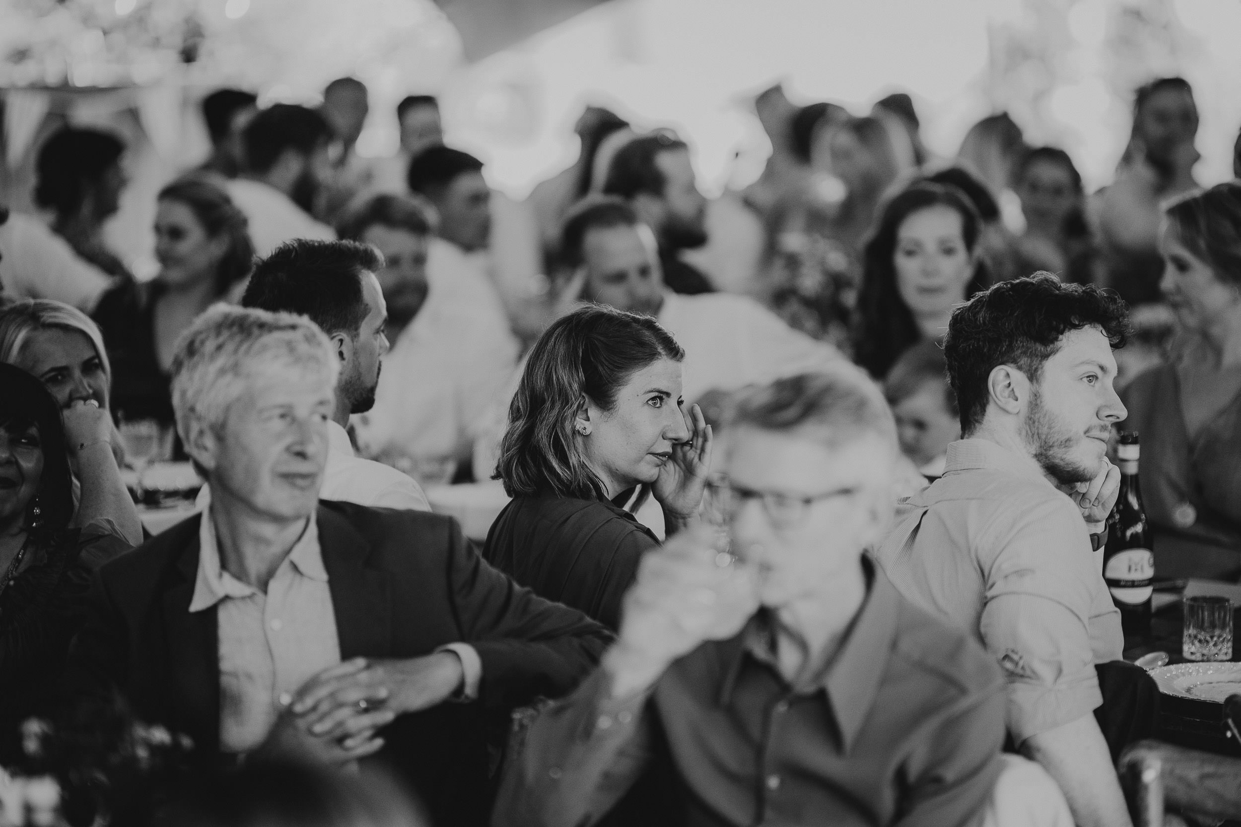 A large group of people seated at tables in a crowded indoor setting, engaged in conversations and listening. The image is in black and white.