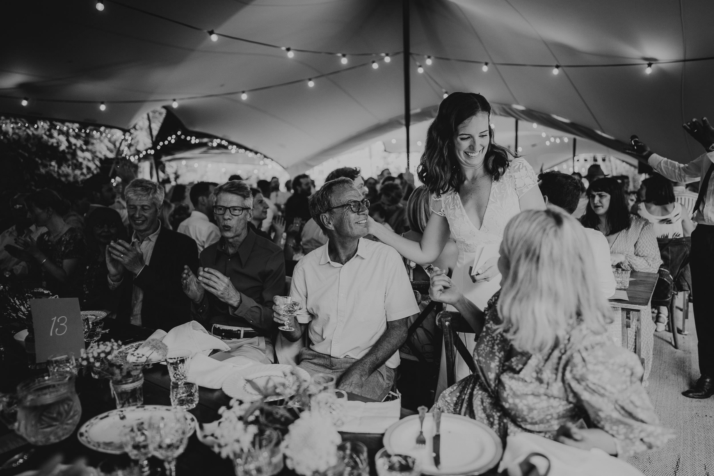 Guests seated at tables under a tent at a wedding reception. A woman in a white dress smiles while interacting with people at a table. String lights are visible overhead.