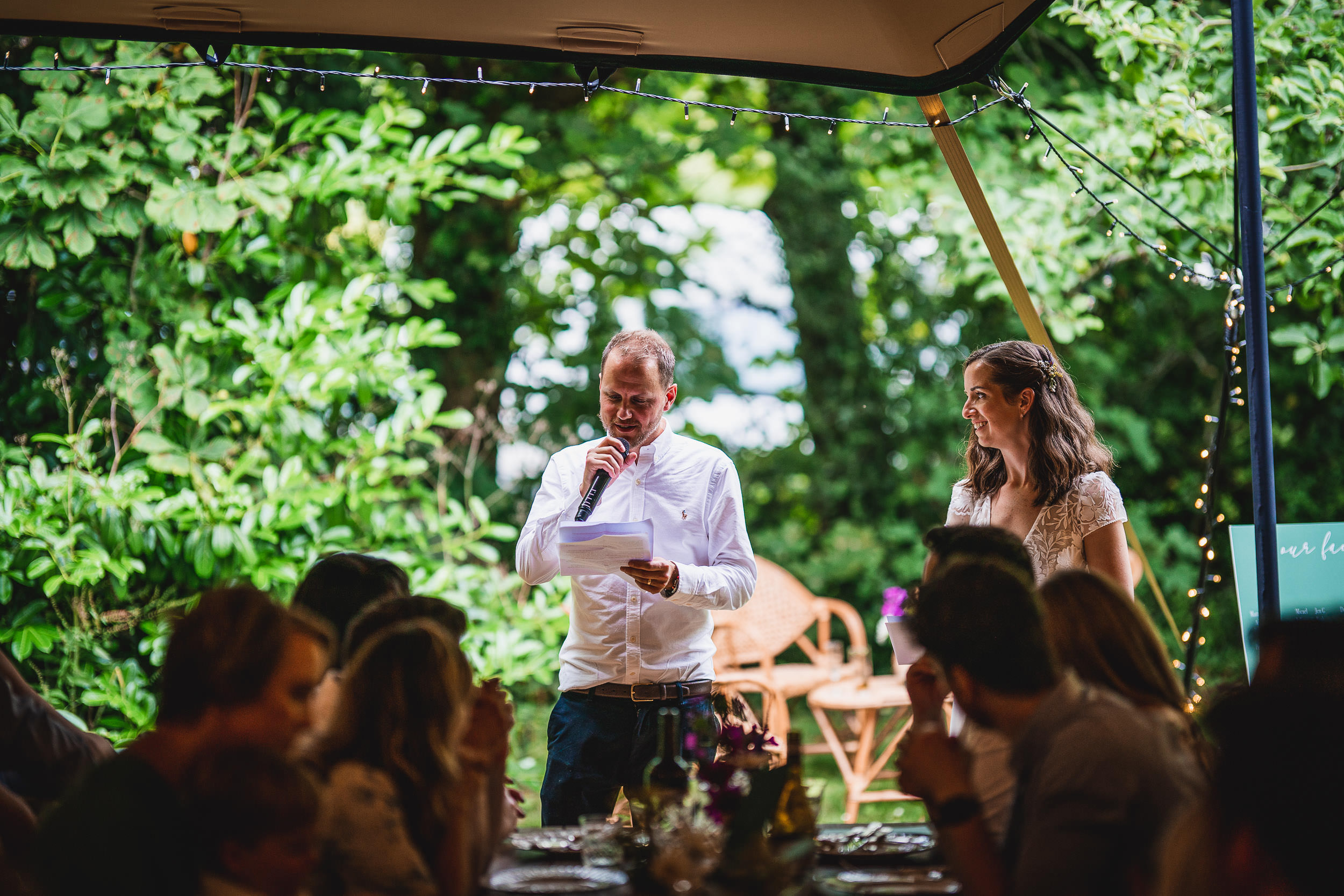 A man and woman stand under a canopy with string lights. The man speaks into a microphone while the woman smiles. Guests sit at tables with blurred greenery in the background.