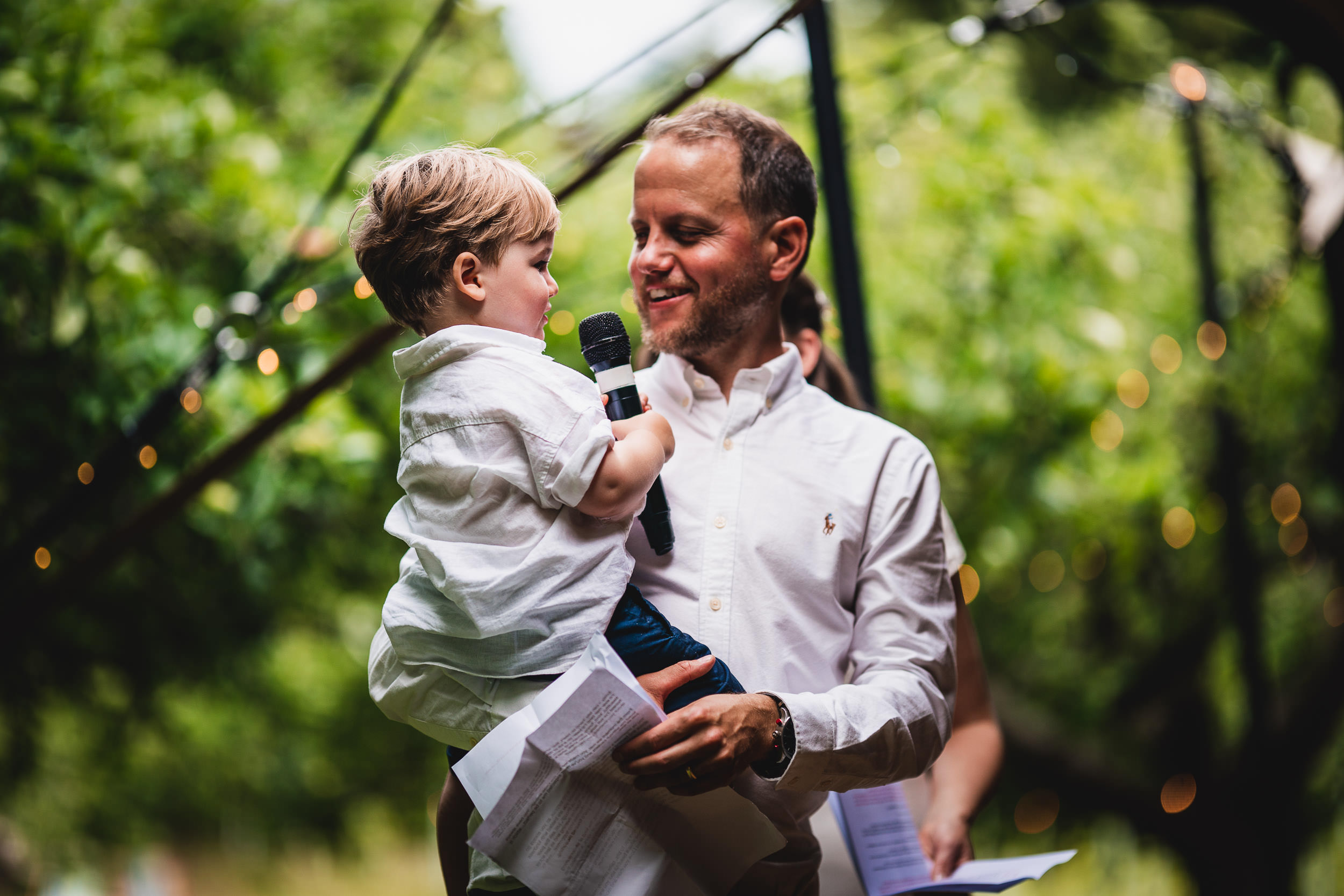 A man holds a child while speaking into a microphone outdoors, surrounded by greenery and festive lights.