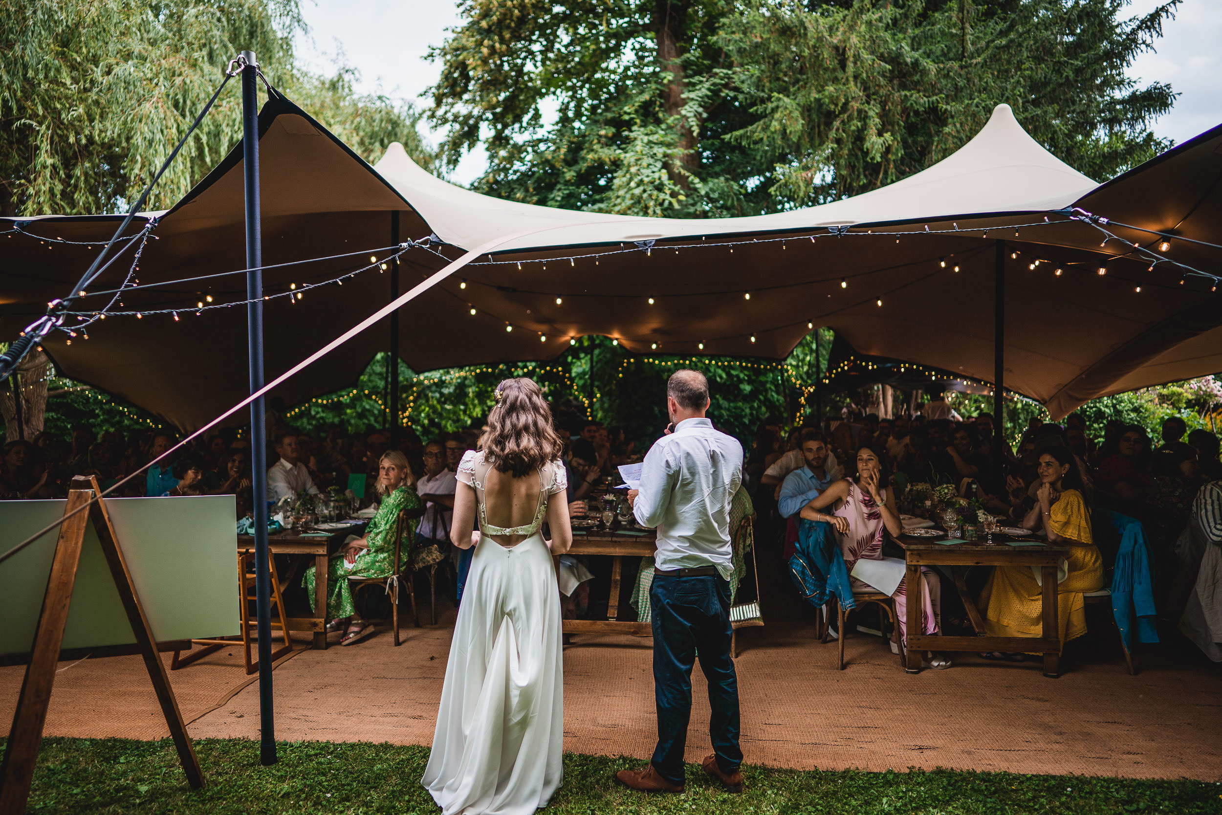 A couple stands at the front of an outdoor gathering under a canopy, addressing guests seated at tables. String lights decorate the tent.