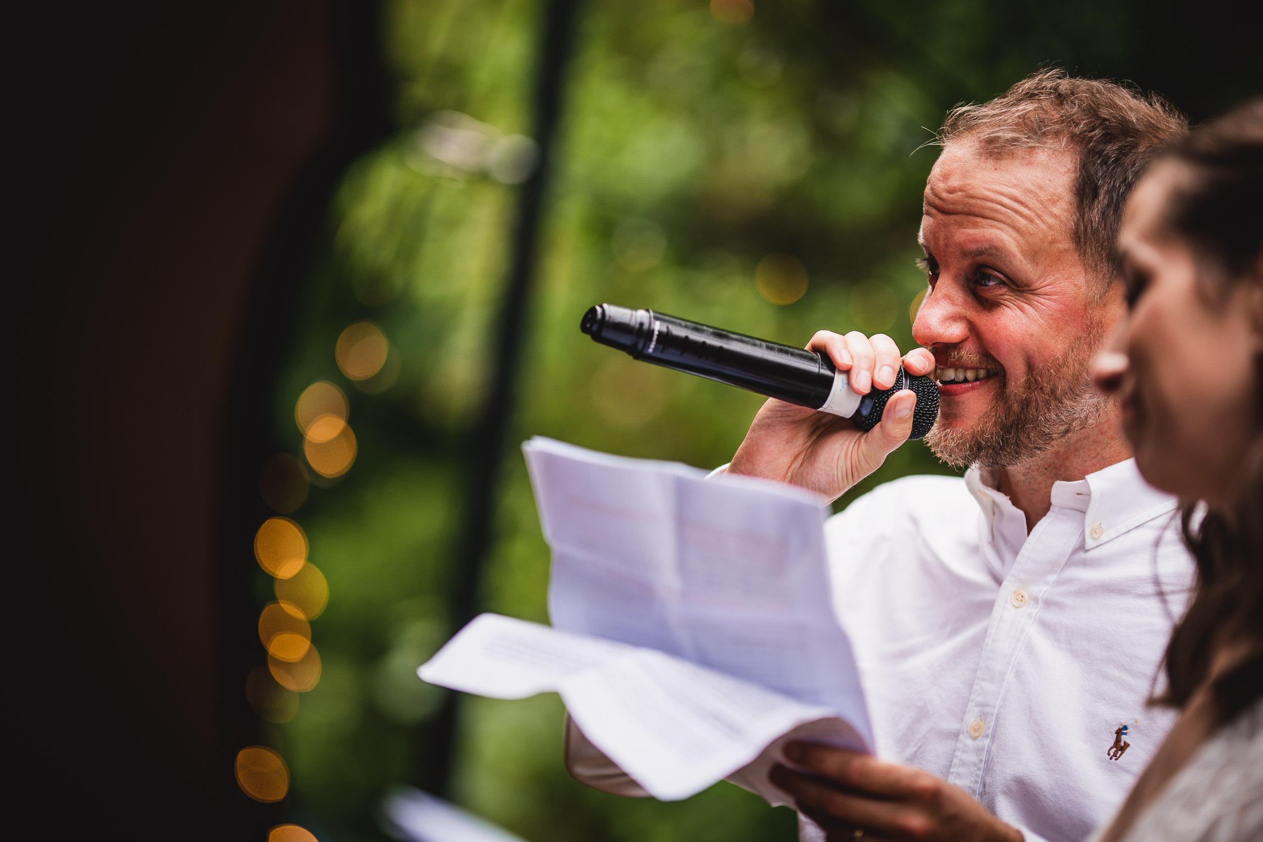 A man in a white shirt holds a microphone and papers, smiling while speaking. A blurred figure stands beside him. Background features soft-focus greenery and twinkling lights.