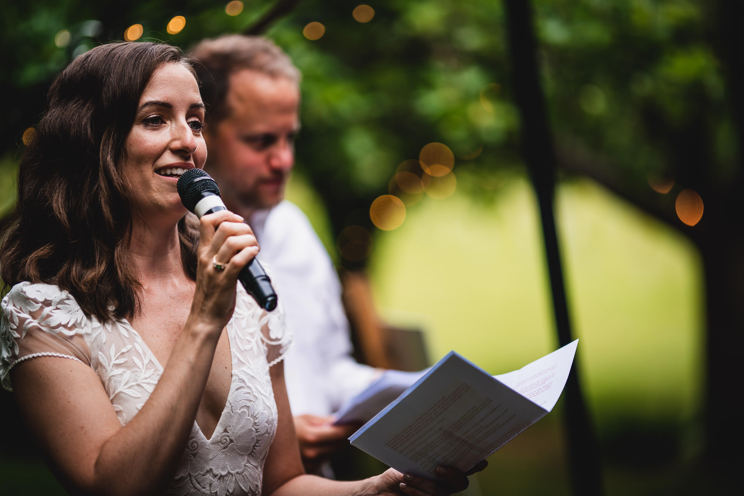 A woman in a white dress is speaking into a microphone, holding papers. A man stands in the background, both outdoors with greenery and lights behind them.