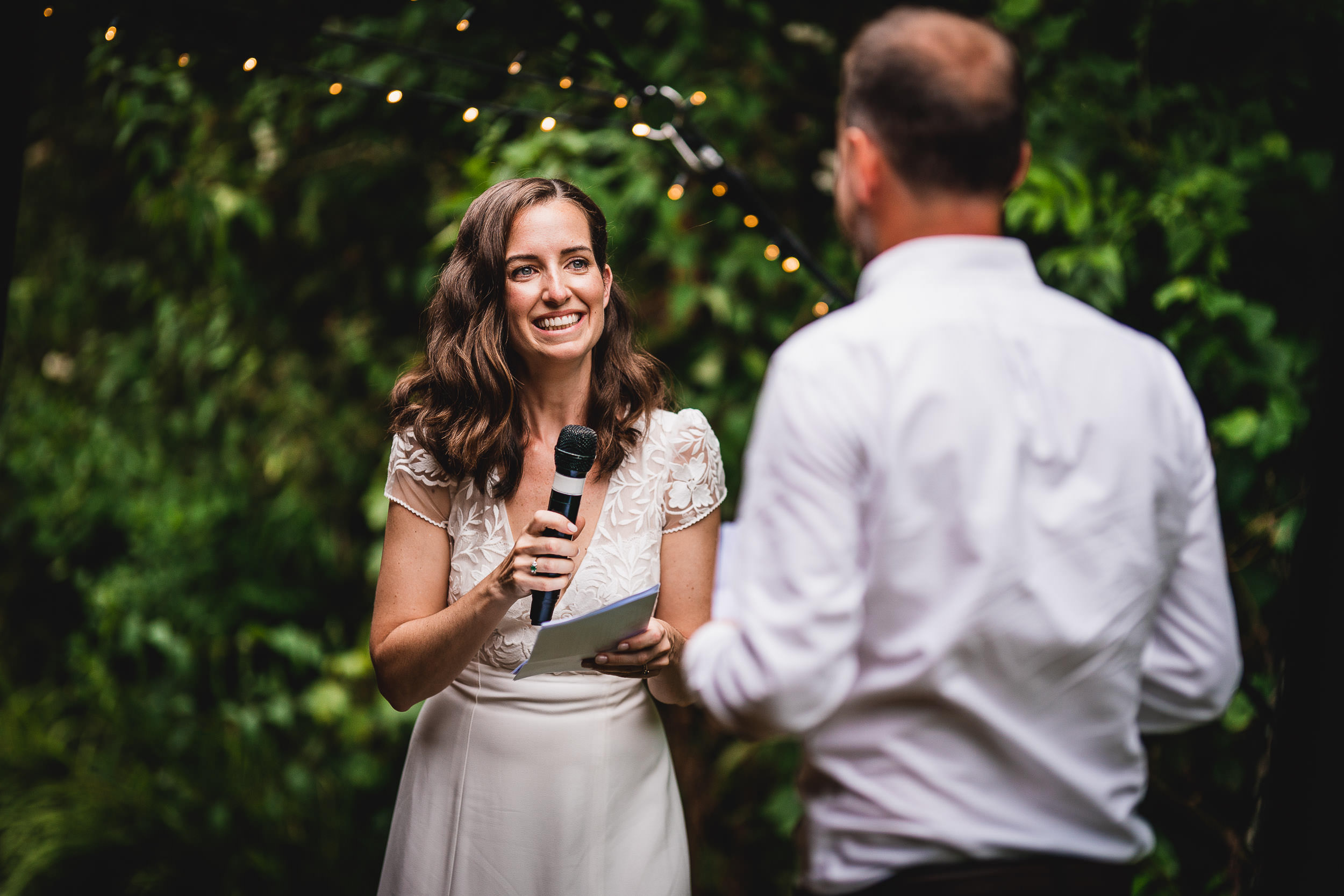 A woman in a white dress holds a microphone and paper, smiling as she faces a man in a white shirt, with greenery and string lights in the background.