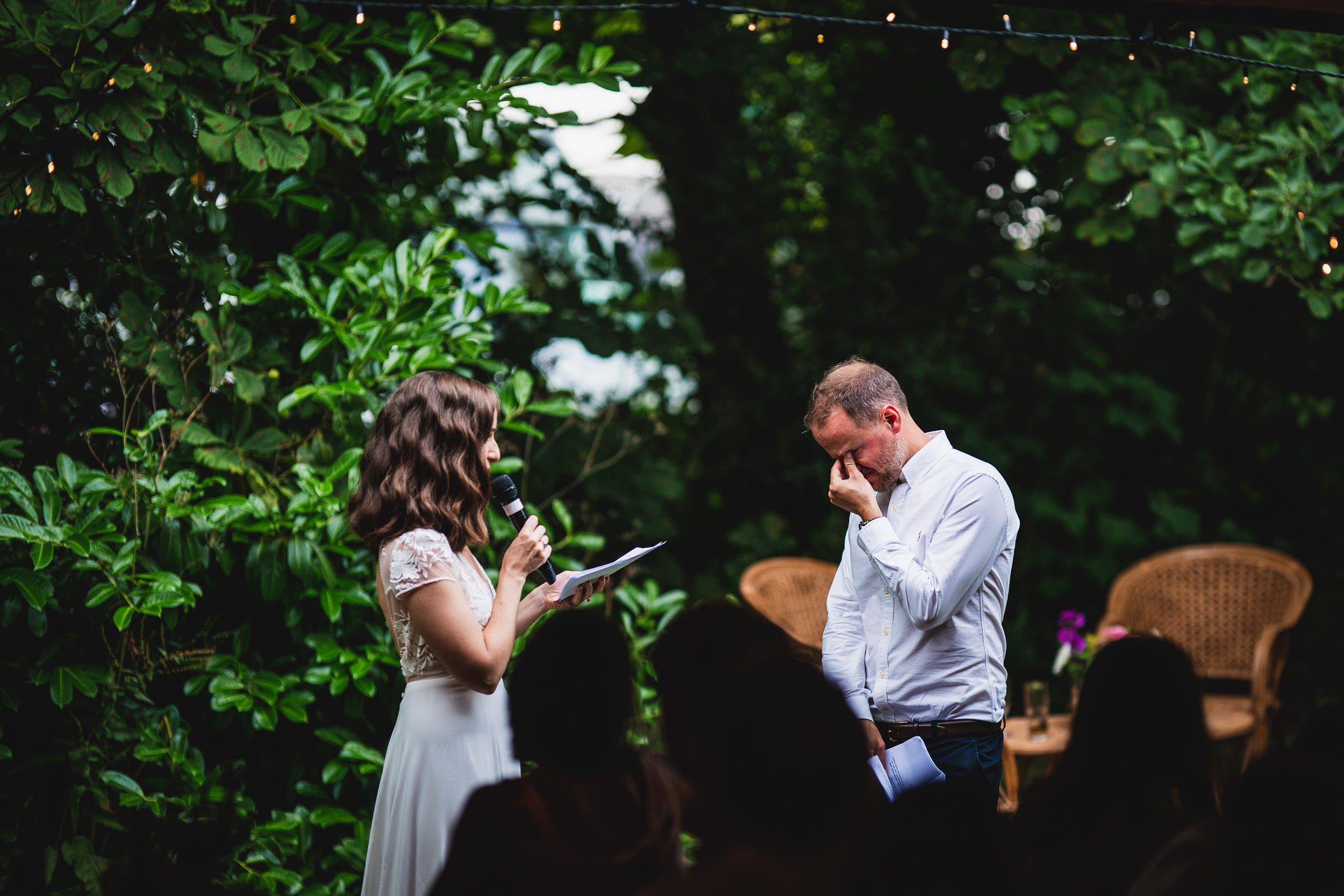 A woman in a white dress speaks into a microphone while a man in a white shirt stands nearby, appearing emotional. They are outdoors, surrounded by lush greenery.