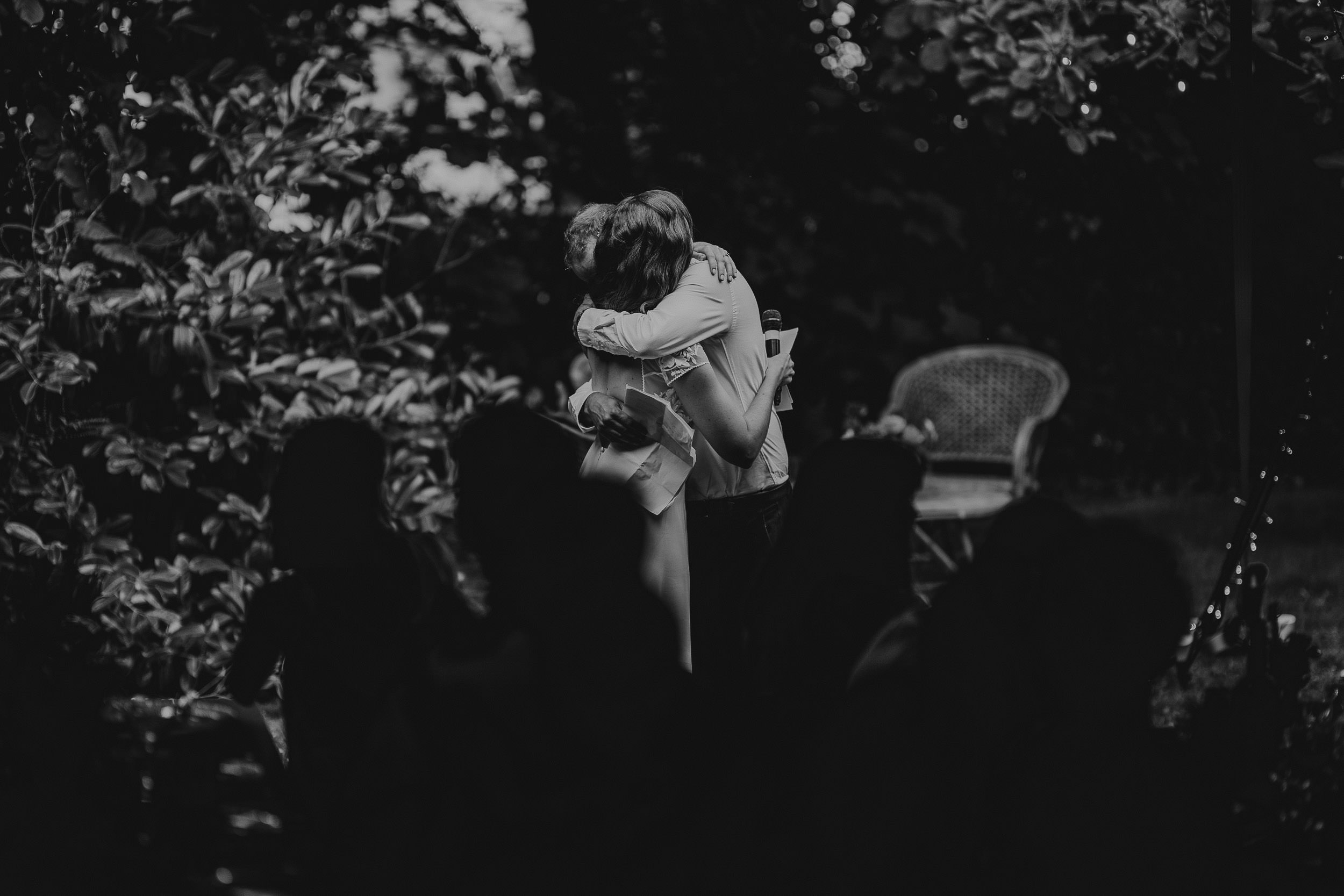 Two people embrace in a garden setting, surrounded by trees and silhouetted figures. A chair is visible in the background. The image is in black and white.