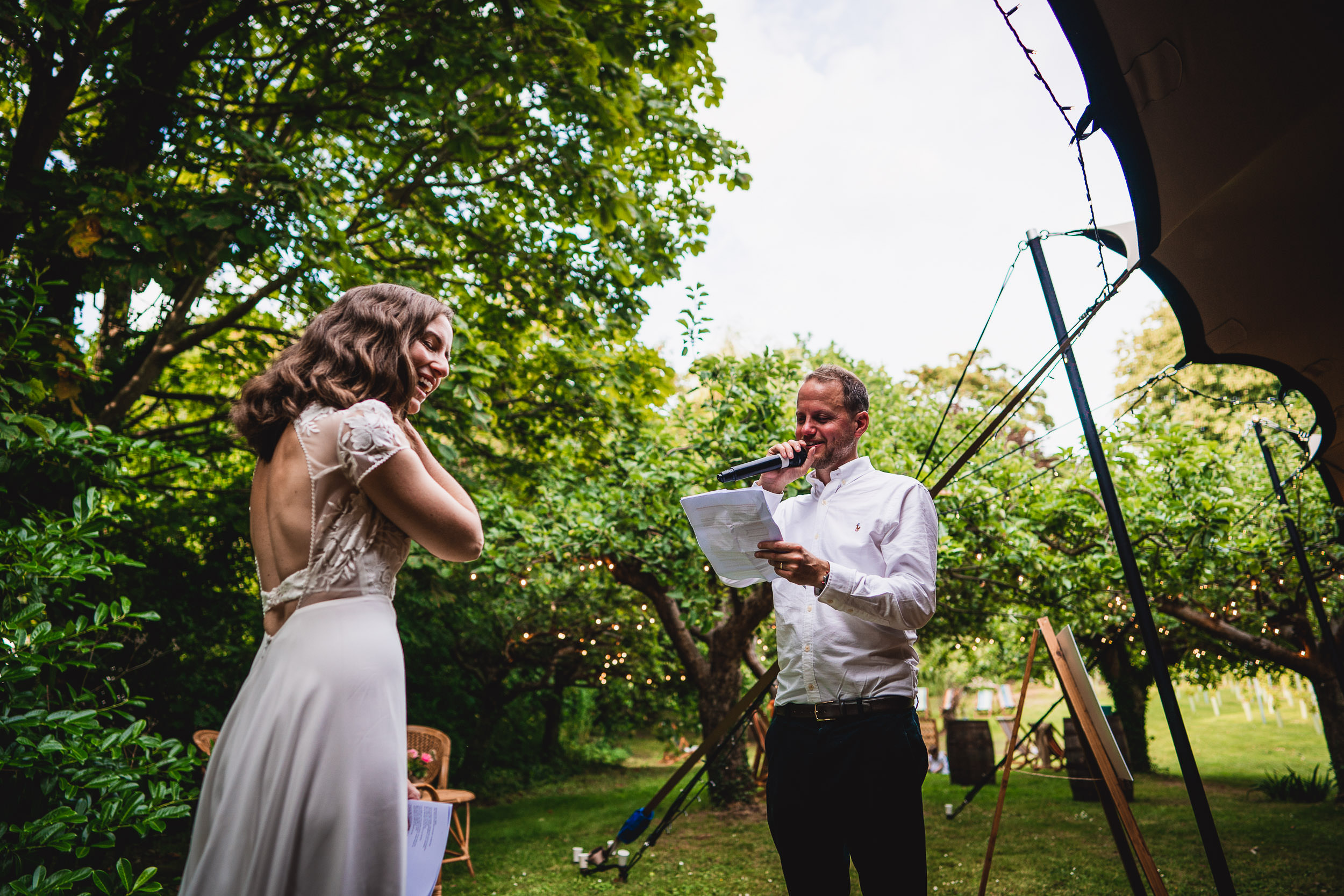 A bride smiles as a man in a white shirt reads from a paper using a microphone, standing in a garden setting with trees and lights.