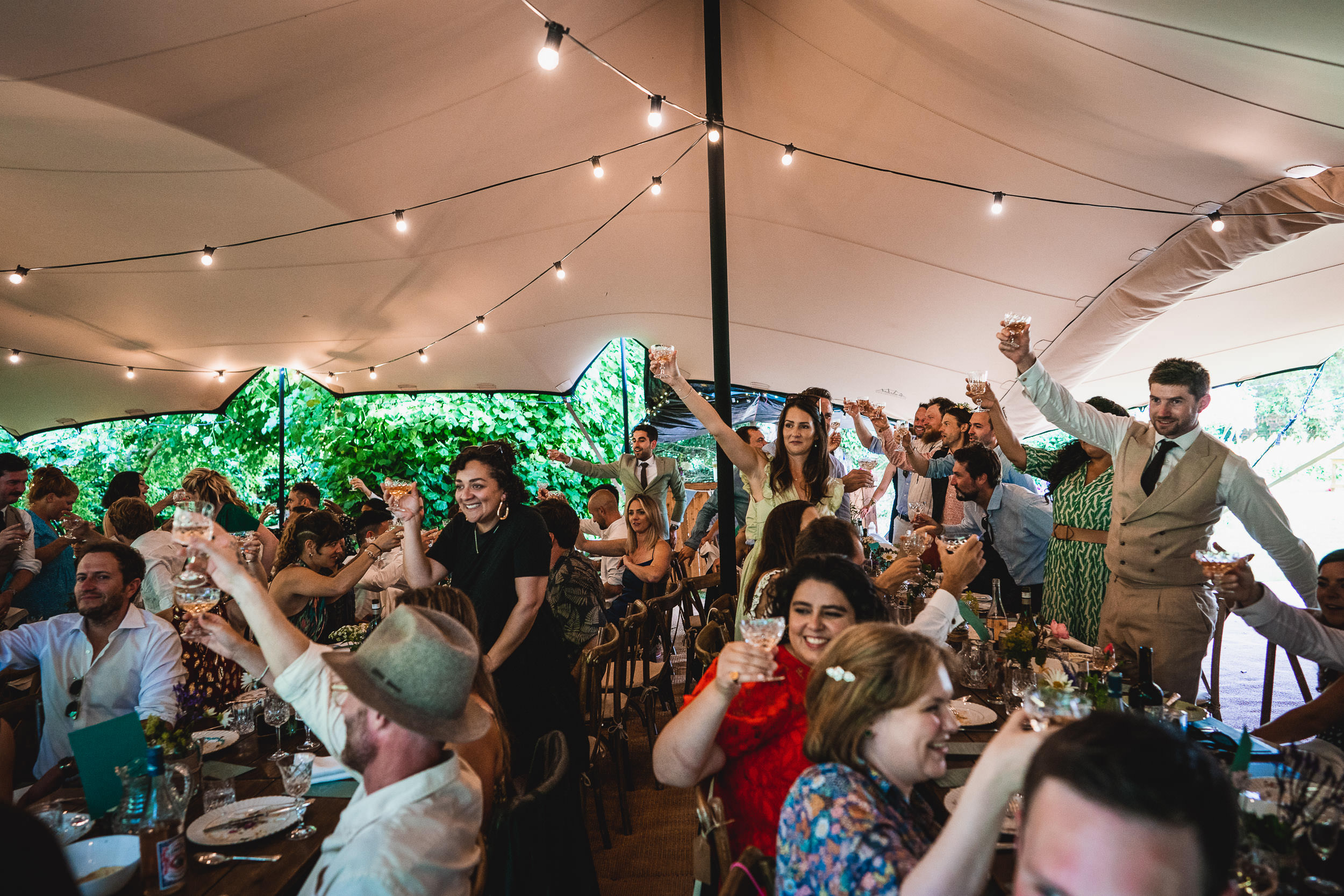 People at an outdoor event under a tent raise glasses in a toast. String lights decorate the space, and greenery is visible in the background.