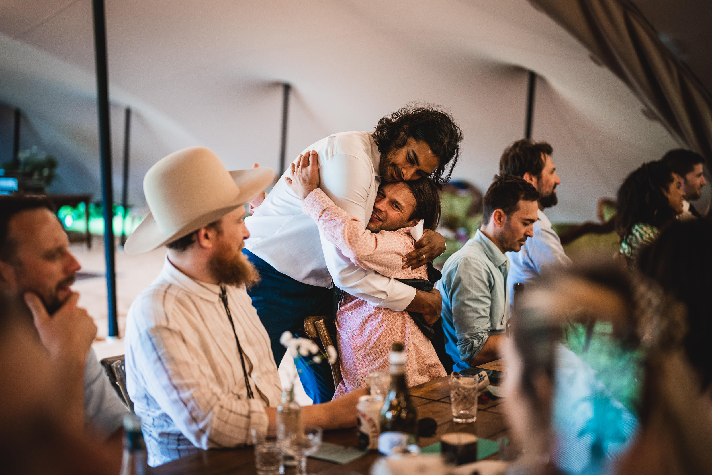 A person embraces another from behind at a dinner event. Several others sit at the table, engaged in conversation.