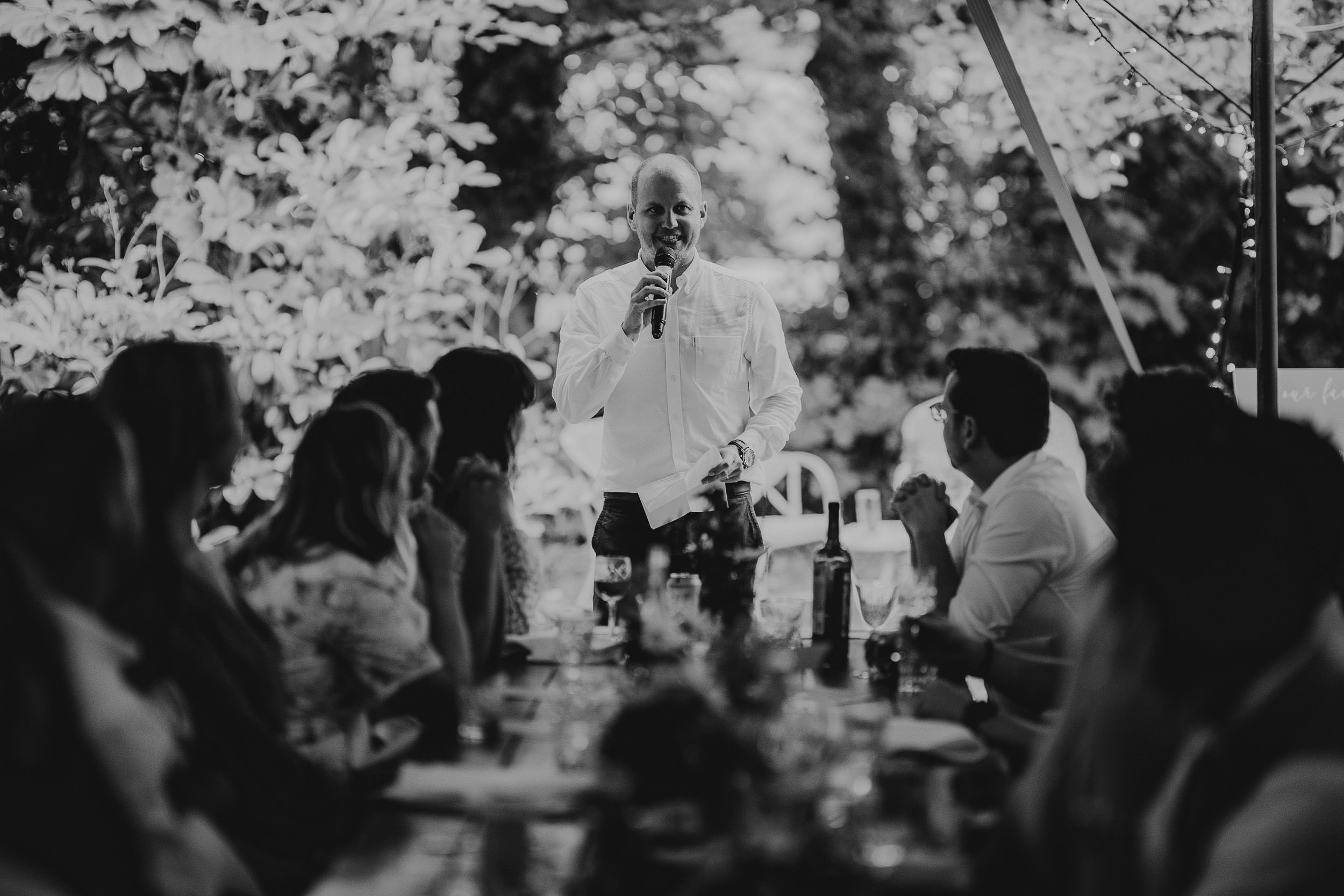 A man stands speaking into a microphone in front of seated people at an outdoor event.