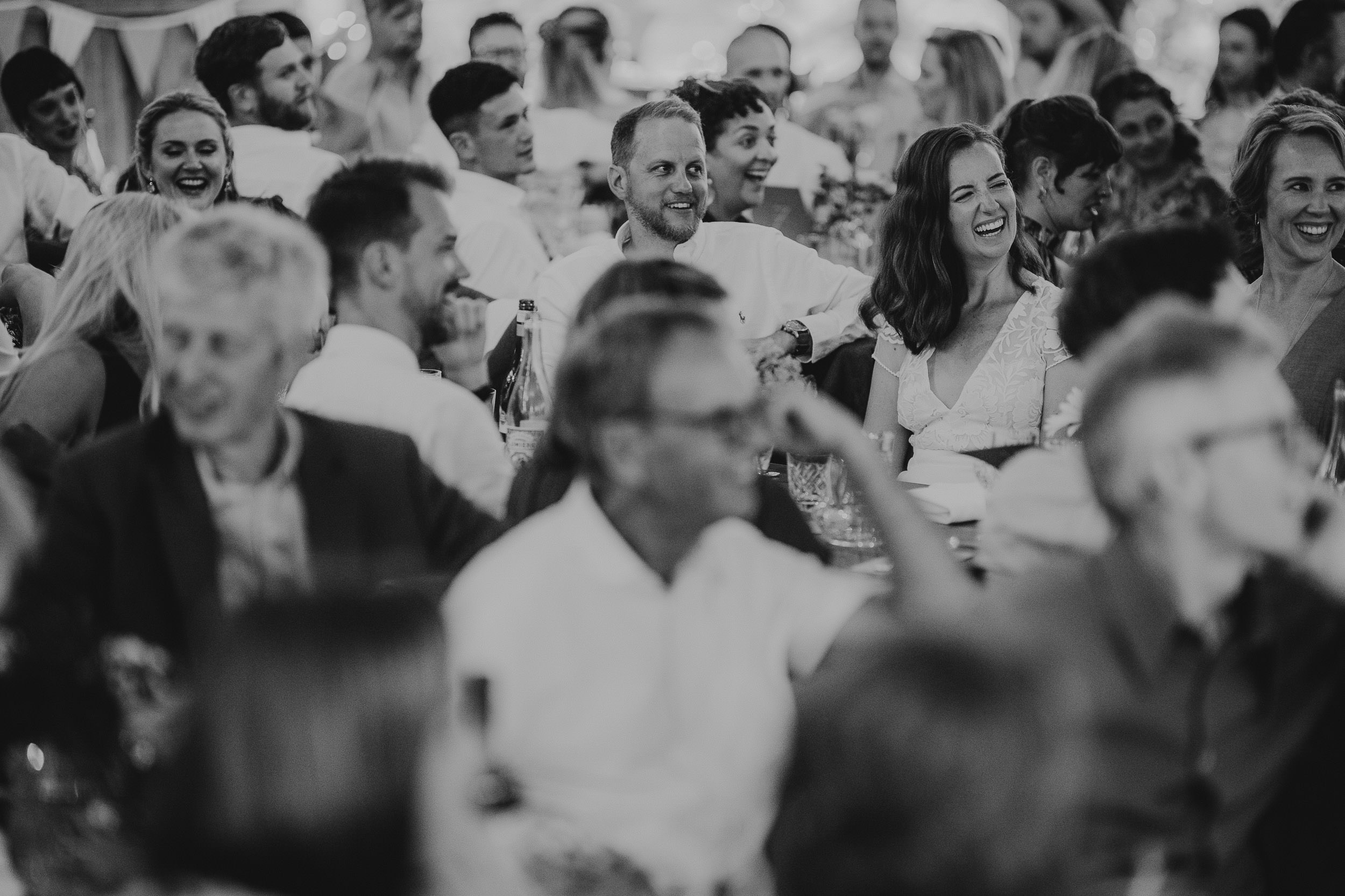 A black and white photo of a group of people sitting at tables, engaged in conversation and laughter at an indoor event.